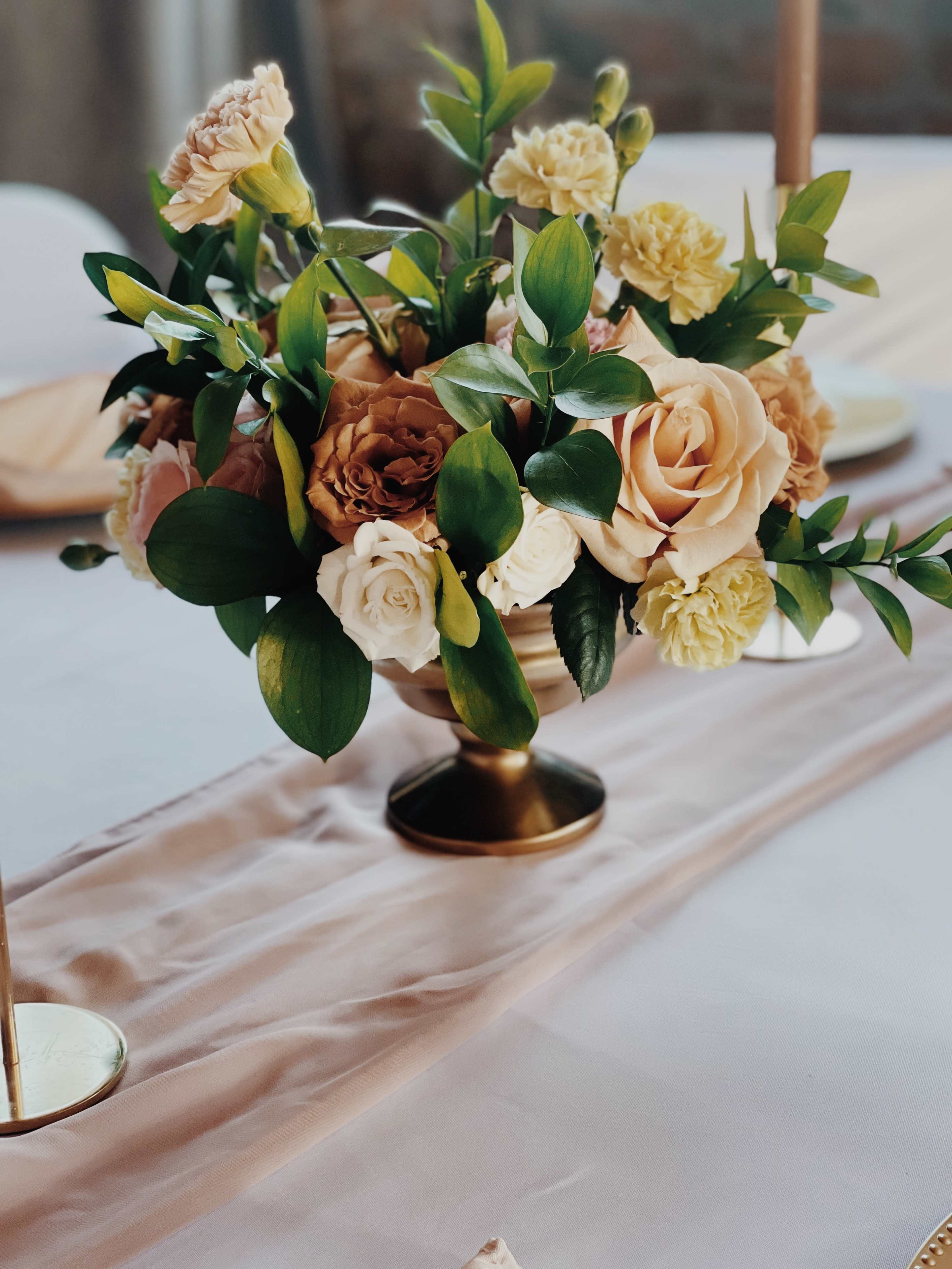 A floral arrangement with roses, carnations, and greenery sits in a gold pedestal vase on a table with a neutral linen runner.