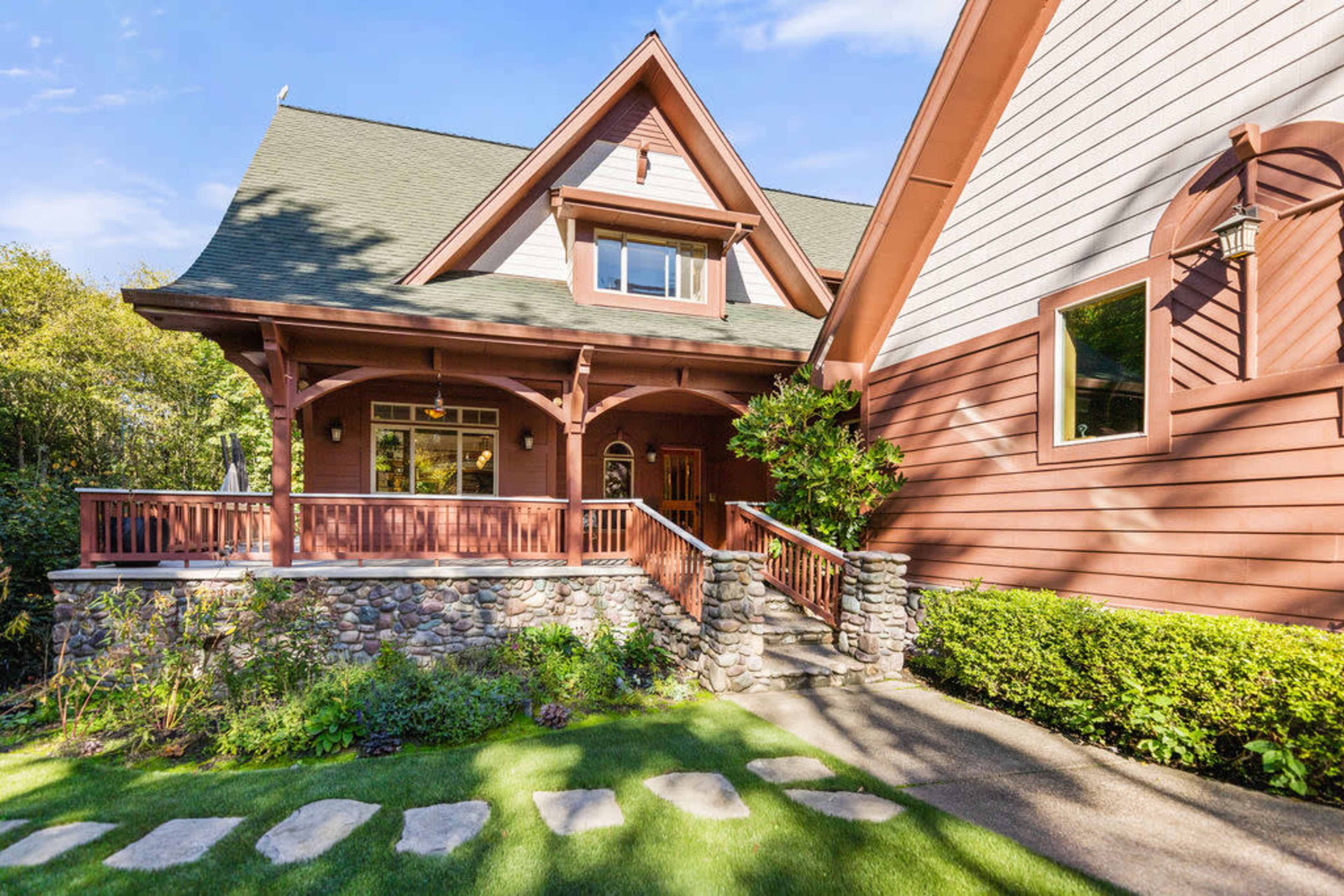 A two-story house with a wooden porch, stone accents, and a sloped roof surrounded by green landscaping.