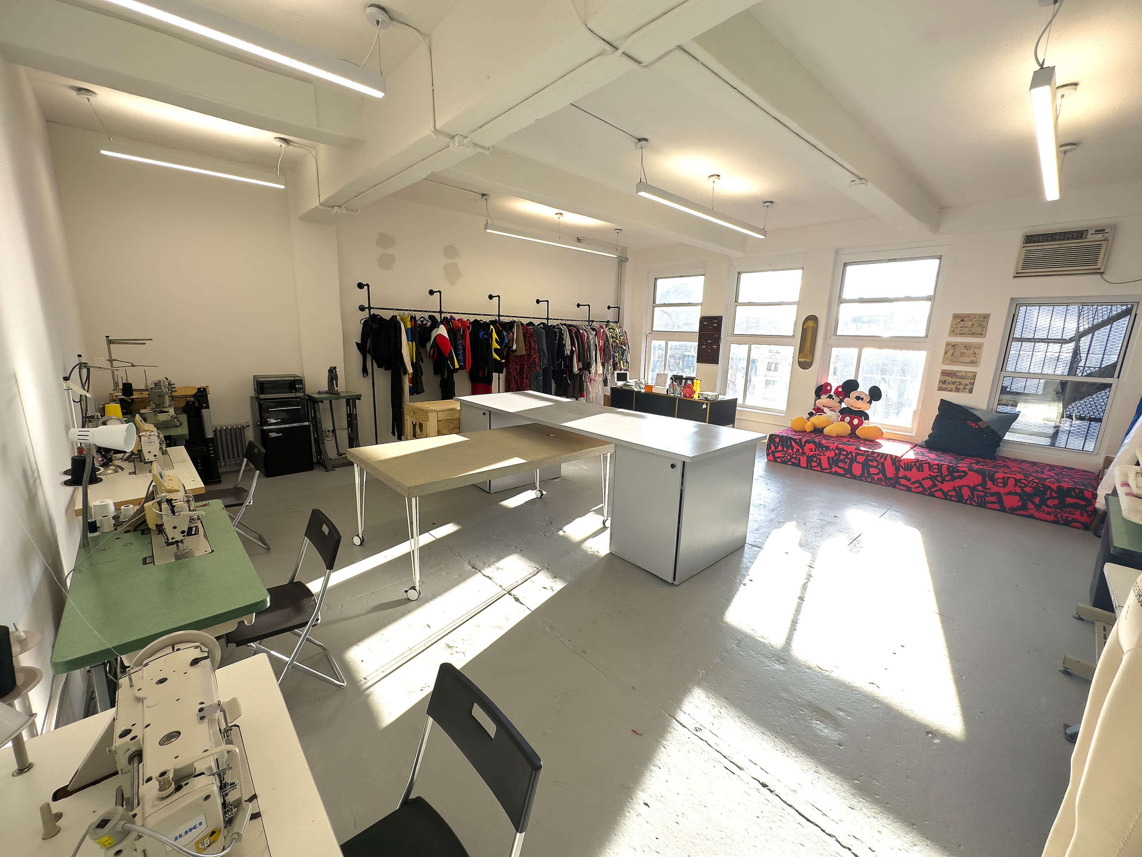 A spacious sewing studio with several machines, work tables, and clothing racks, illuminated by natural light from large windows.
