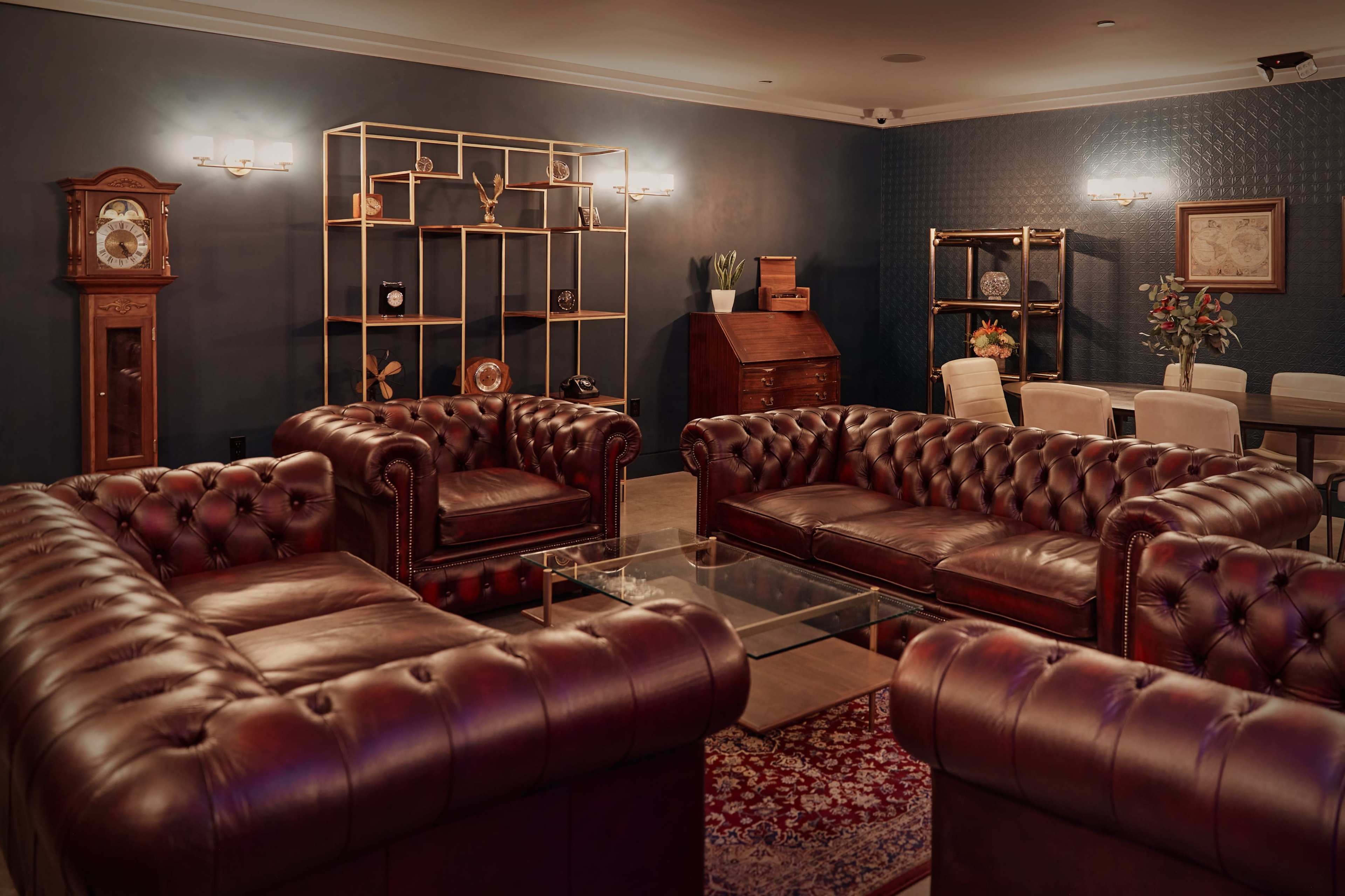 A stylish, dark-hued lounge area featuring multiple brown leather chesterfield sofas arranged around a glass coffee table, with decorative shelves and a clock on the wall.