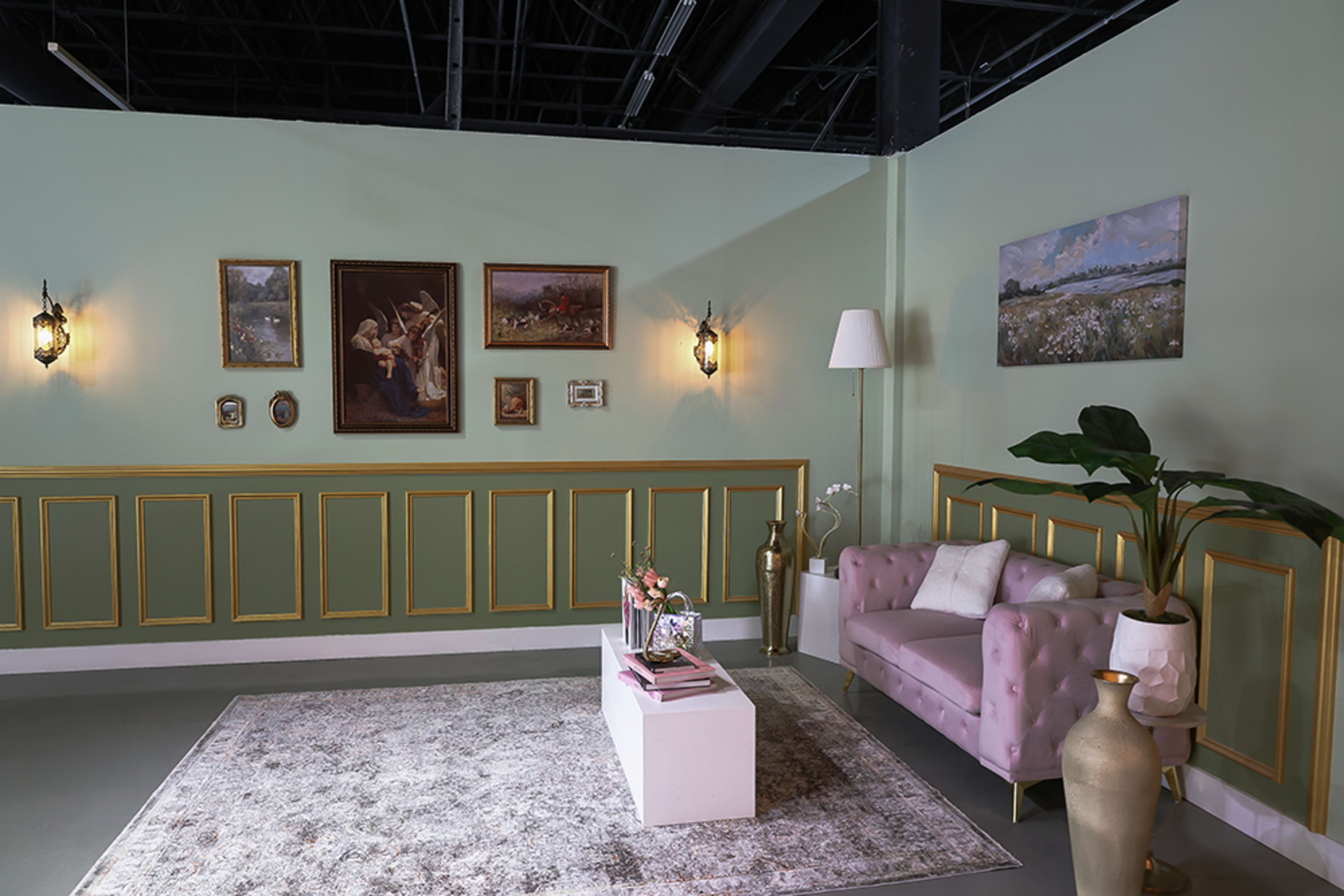 The image shows a stylish interior with green walls, framed artworks, a pink tufted sofa, a white coffee table, and a decorative plant in a corner.
