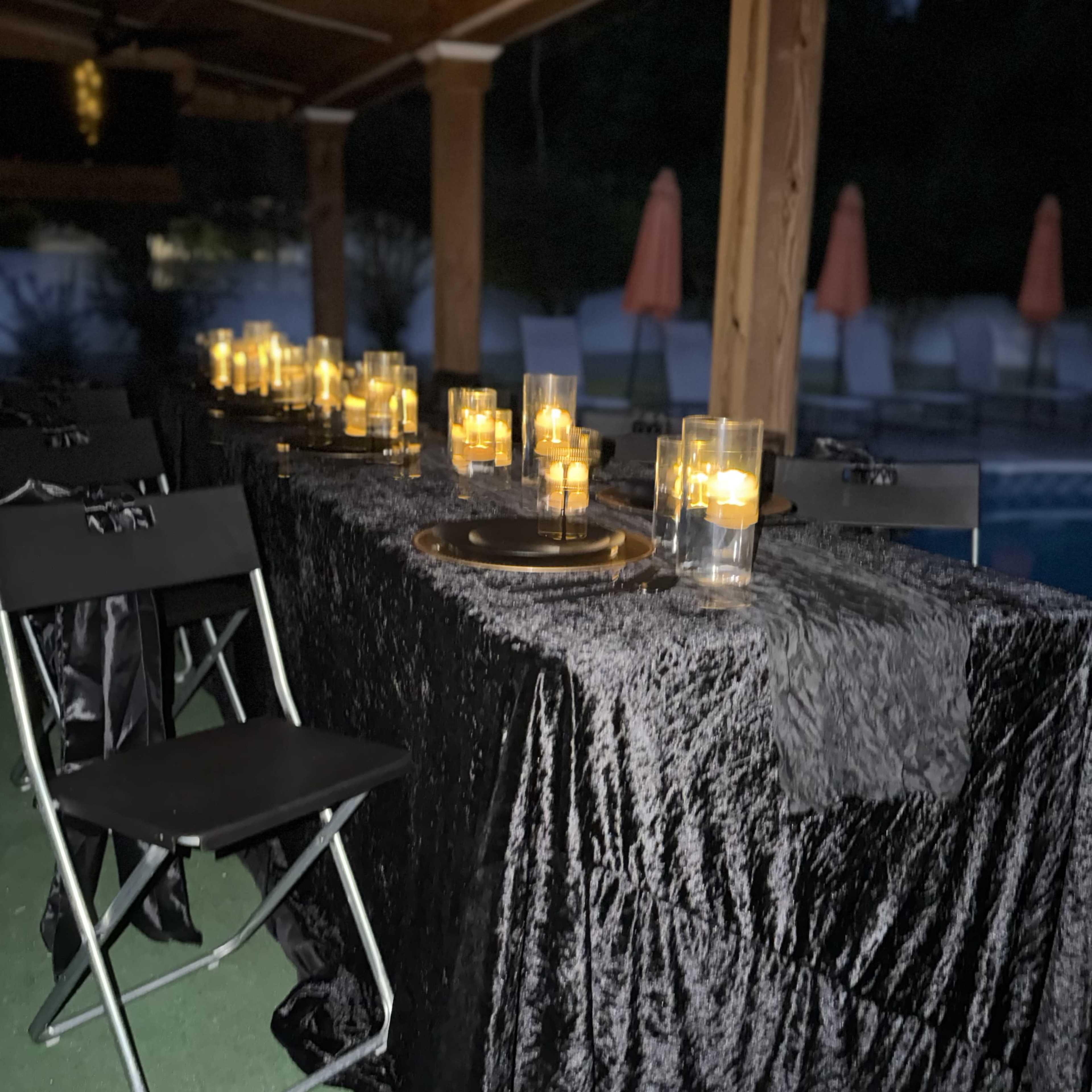 A long table decorated with black cloth and illuminated by candles stands beside a pool at night.