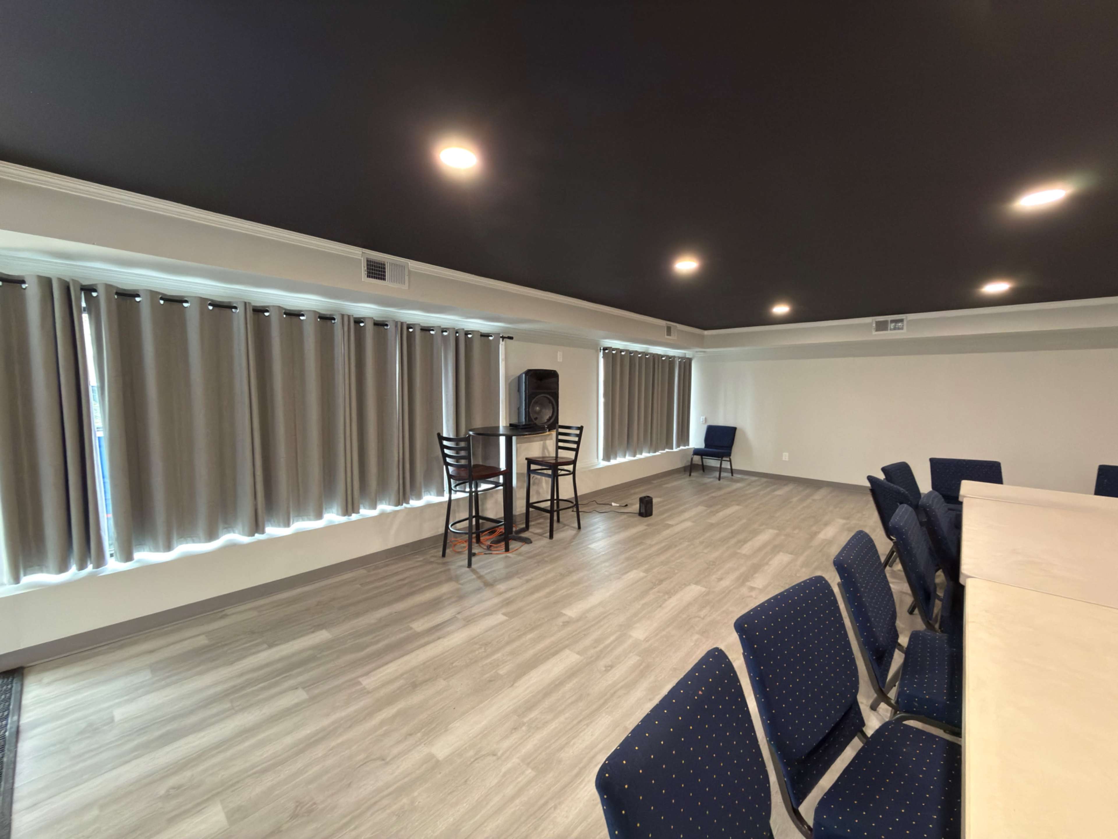 The image shows a spacious, well-lit room with tables, chairs, and large windows covered by curtains.