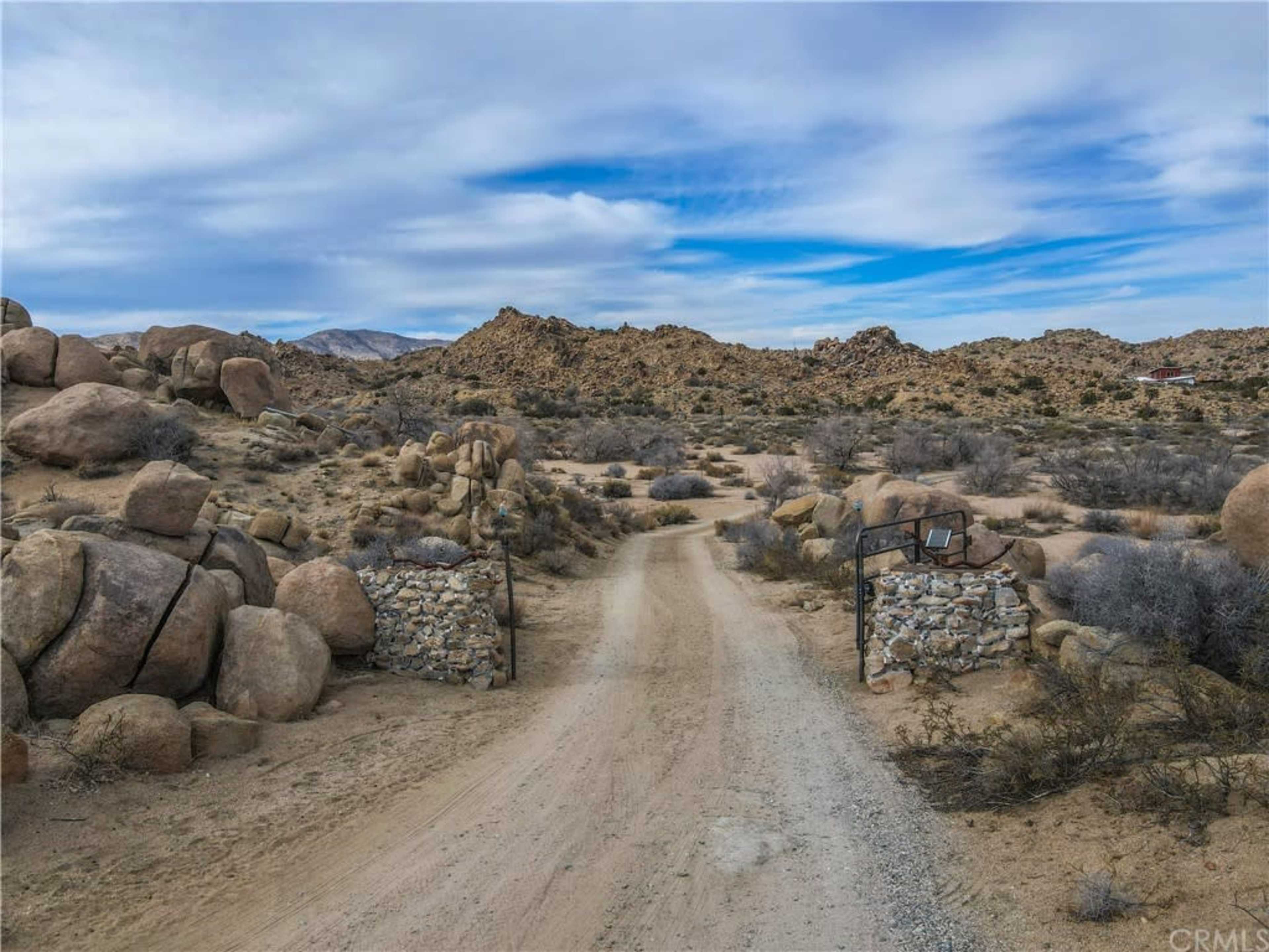 A dirt road leads through stone gate pillars into a desert landscape with rocky terrain and sparse vegetation under a cloudy sky.