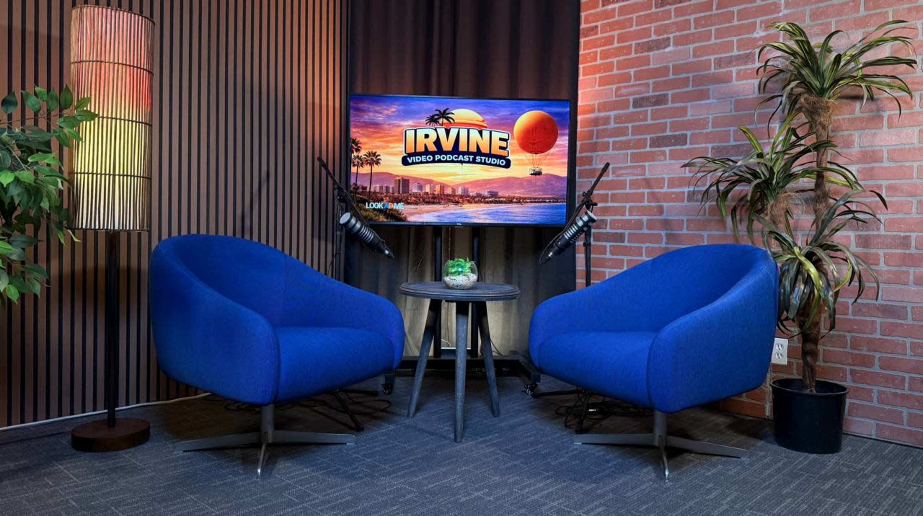The image shows a podcast studio setup featuring two blue chairs, a round table with a plant, a television screen displaying "IRVINE VIDEO PODCAST STUDIO," and a brick wall background.