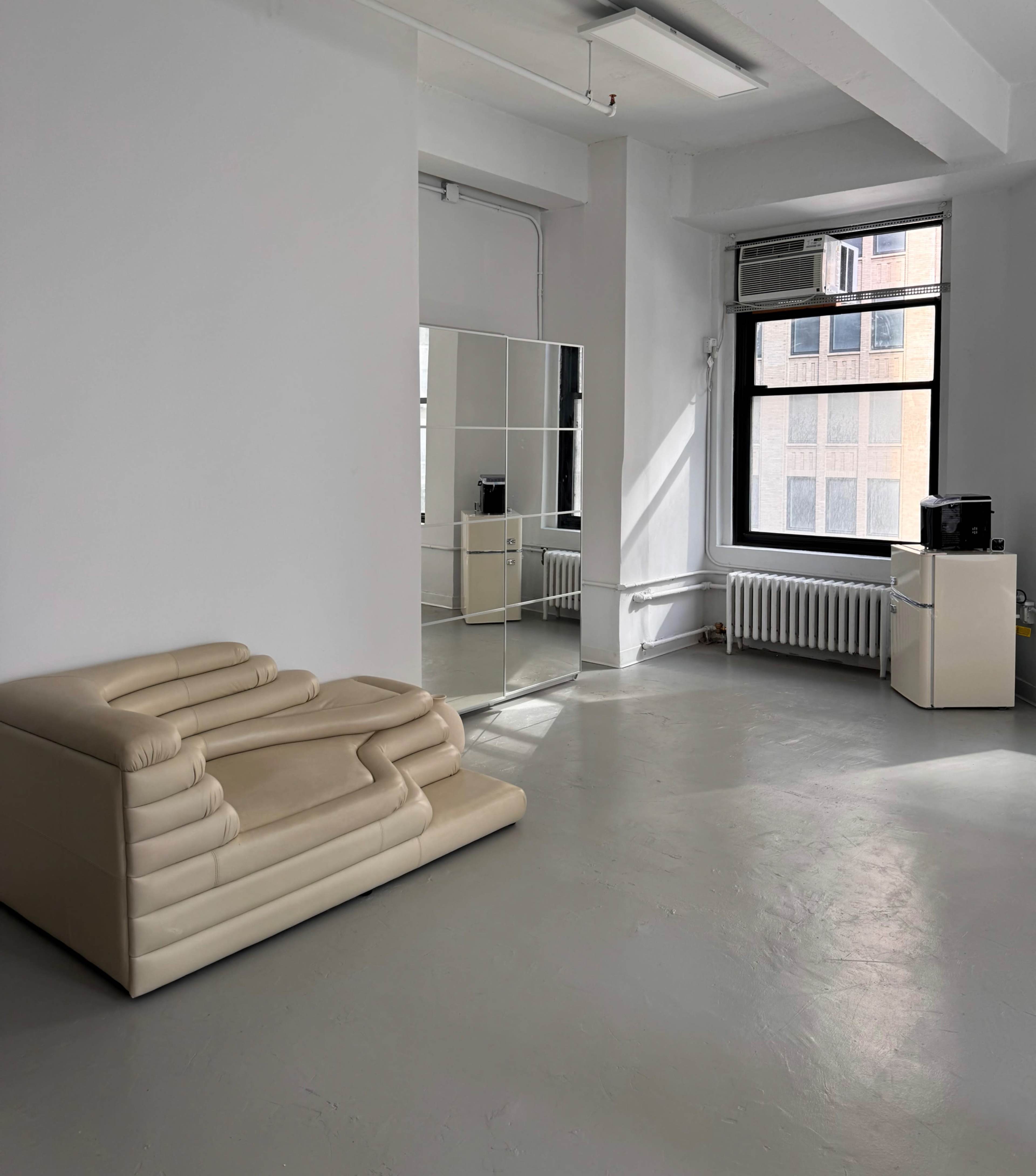 The image shows a minimalist room with a beige sofa, a large mirror, a window with air conditioning, and a small black television on a white cabinet.