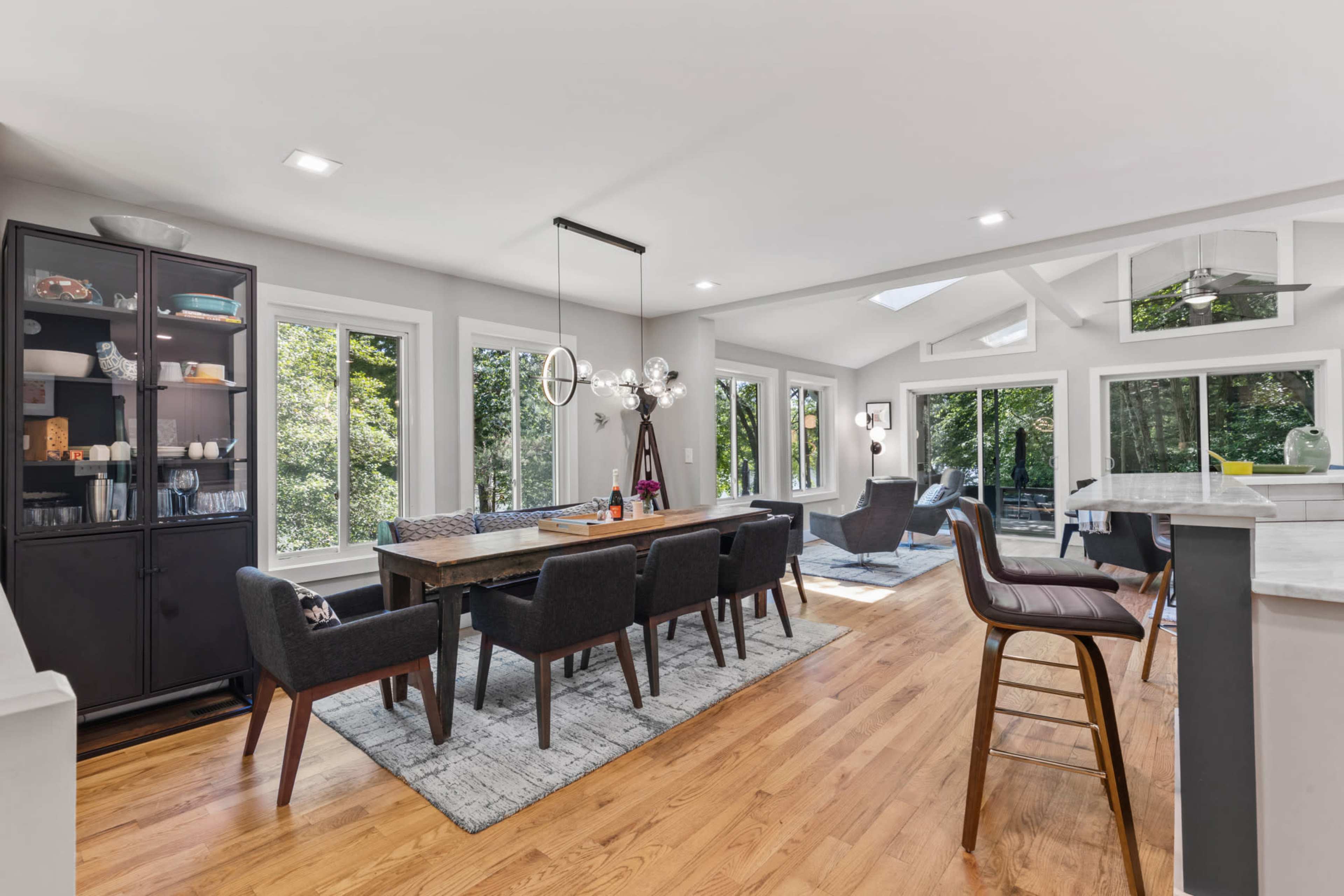 The image shows a modern dining area with a long wooden table surrounded by black chairs, large windows providing natural light, and a sleek kitchen bar in the background.