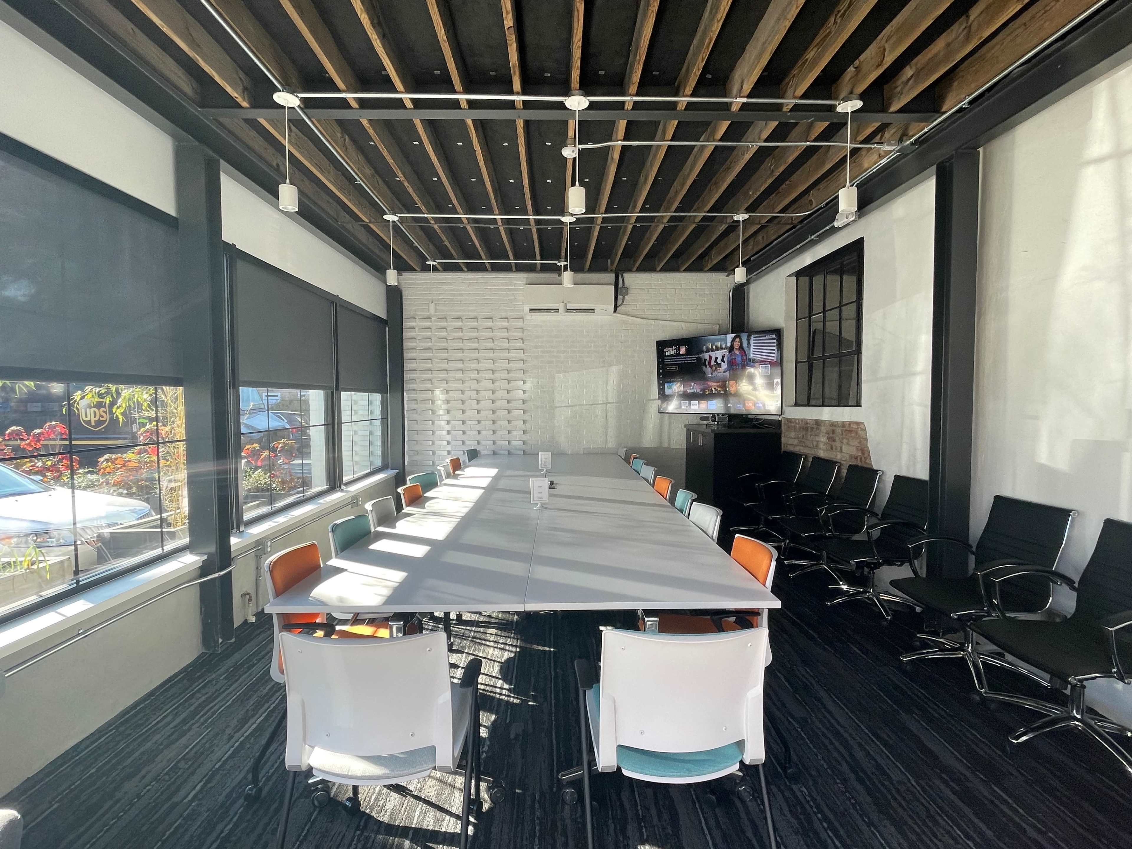 A large meeting room features a long table surrounded by black and orange chairs, with windows that provide views of an outdoor area and a television mounted on the wall.
