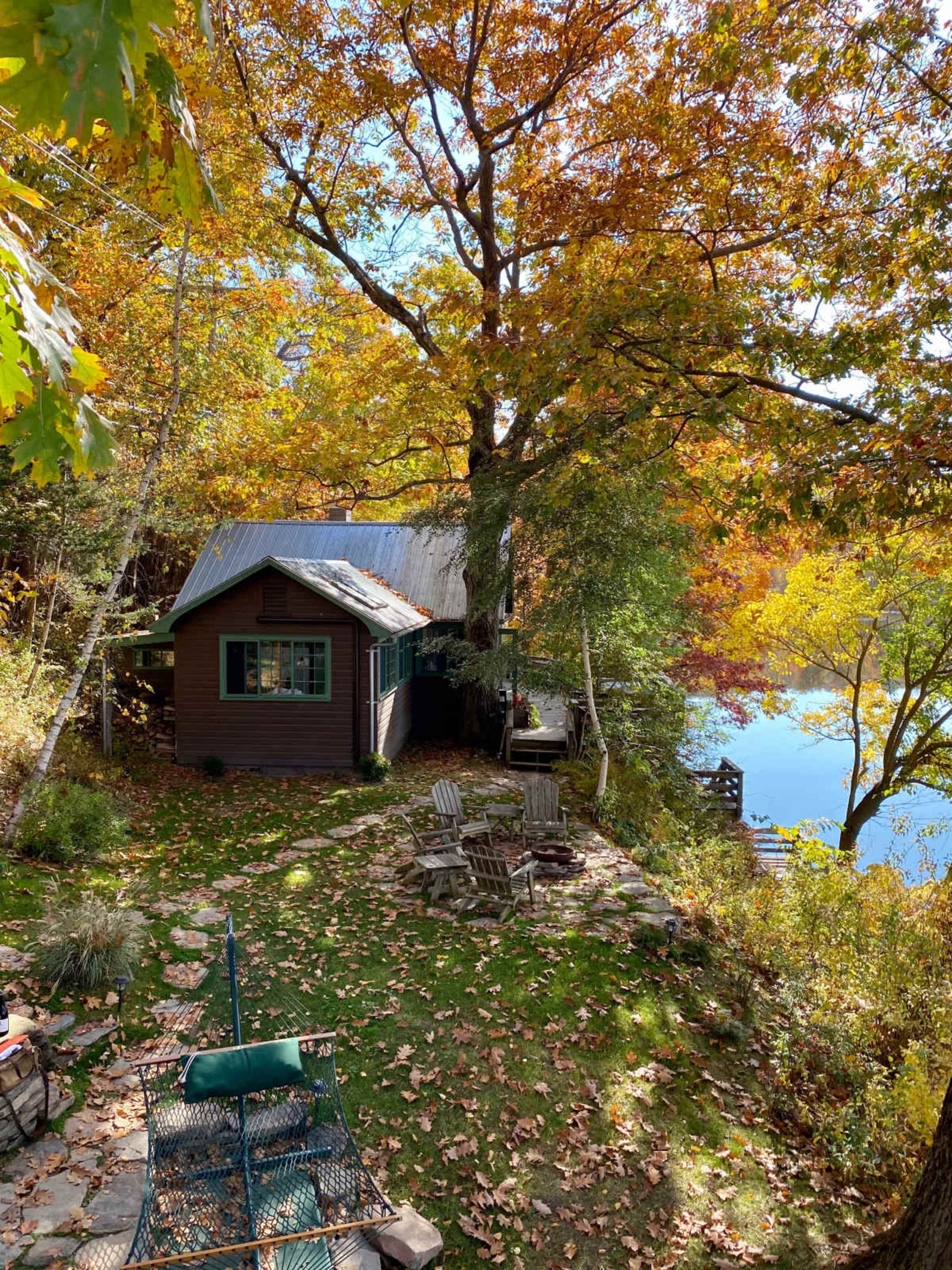 A rustic cabin surrounded by vibrant autumn foliage sits beside a calm lake, with seating areas made of stone and wood.