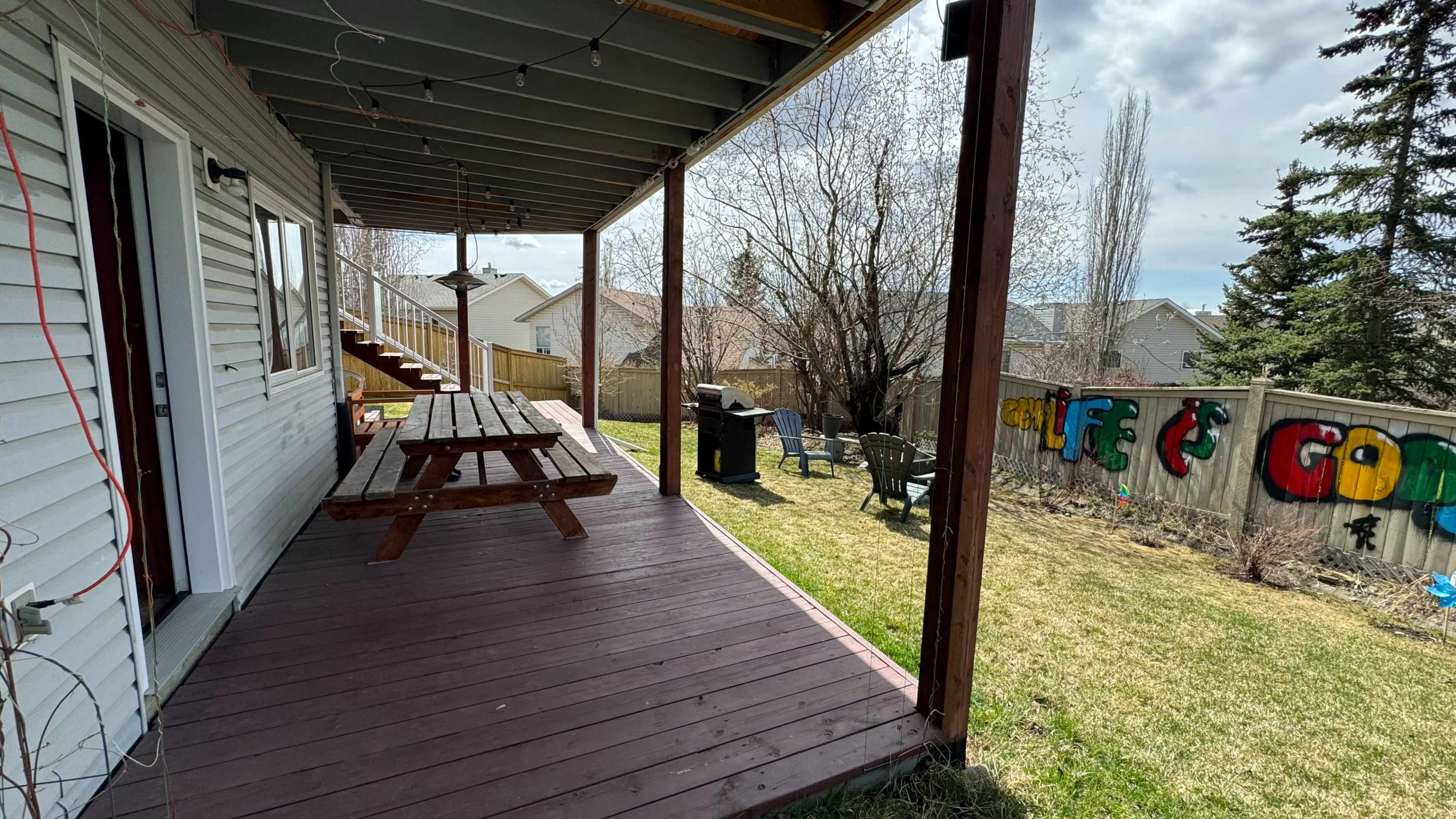 A wooden deck with a picnic table and grill overlooks a backyard featuring a grassy area and a fence with colorful graffiti.