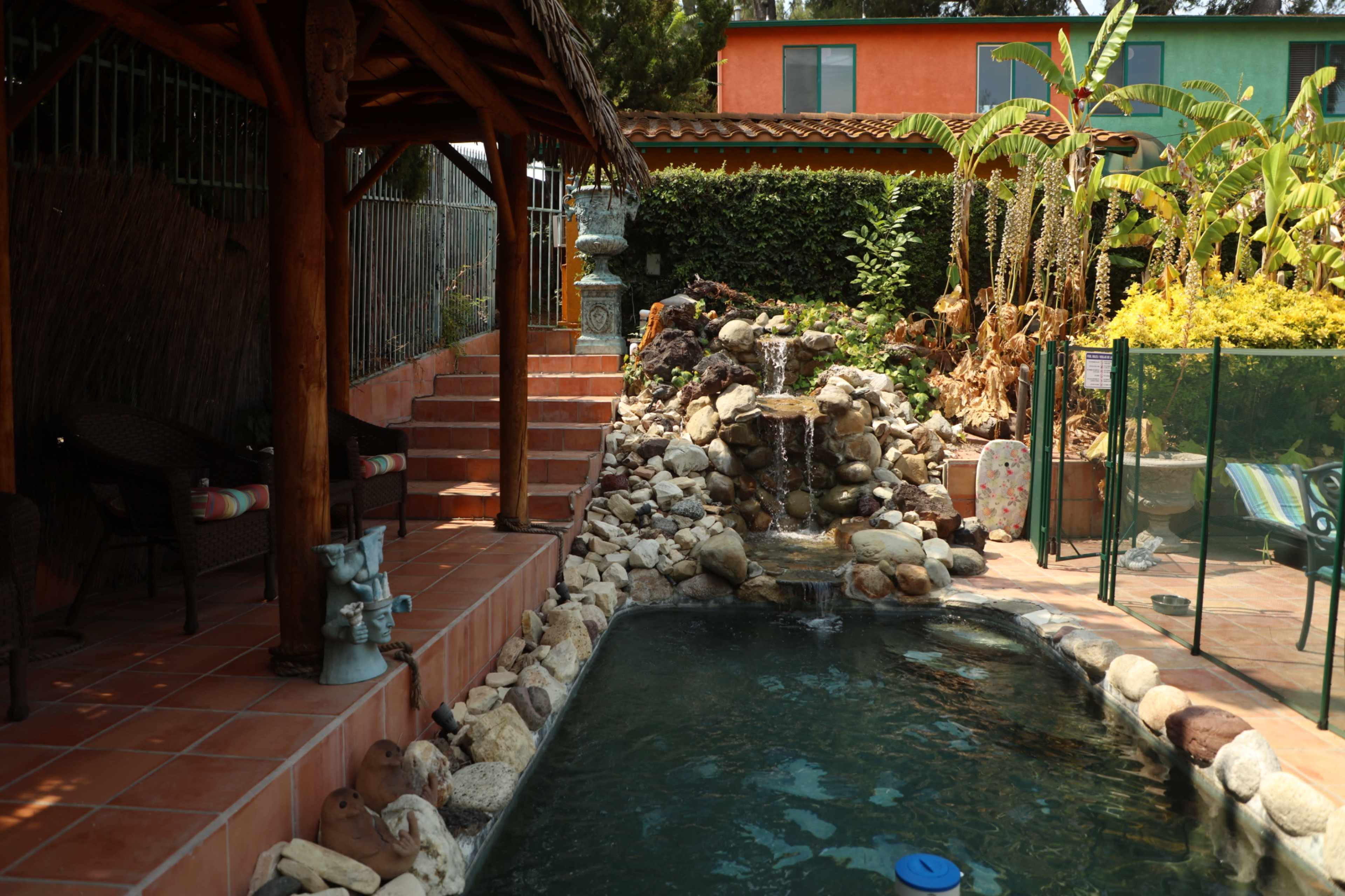 A landscaped backyard featuring a stone fountain beside a swimming pool with surrounding stones and a thatched roof gazebo.