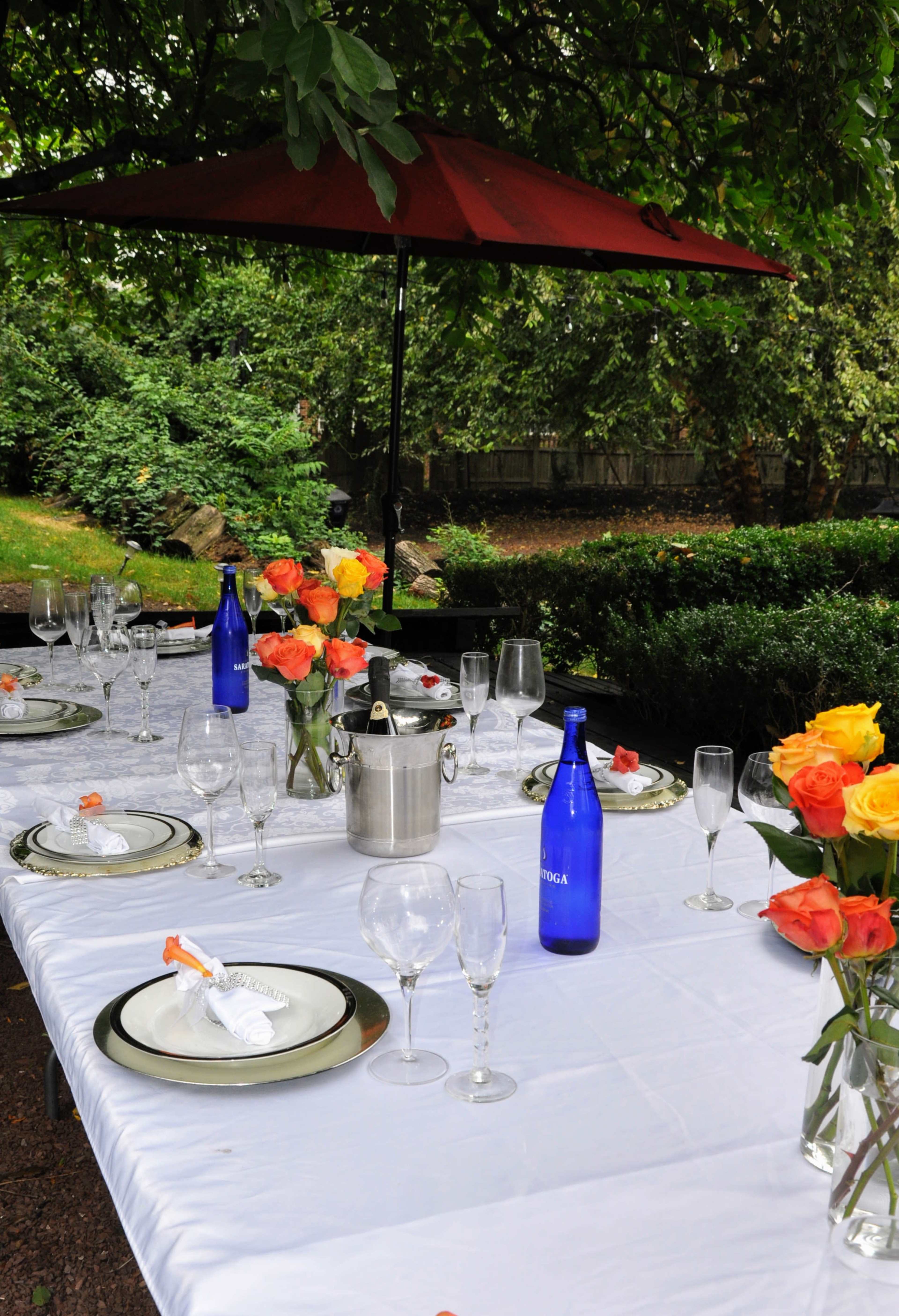 A dining table is set outdoors under a red umbrella, decorated with floral arrangements and accompanied by glasses and bottles of water.