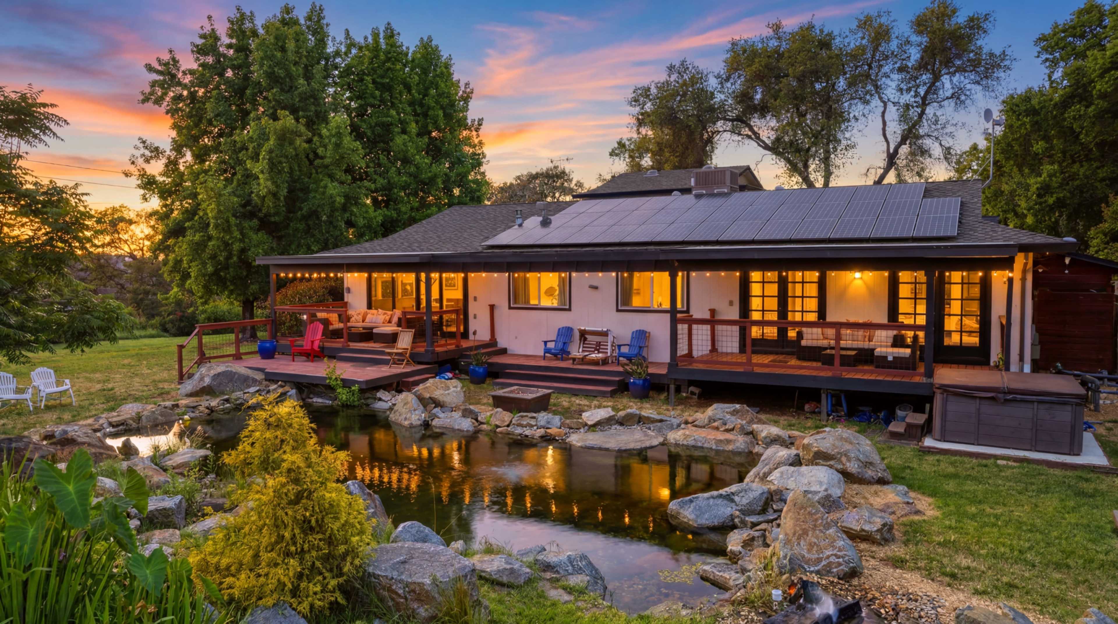 A modern house with solar panels sits by a pond surrounded by rocks and greenery, illuminated by sunset.
