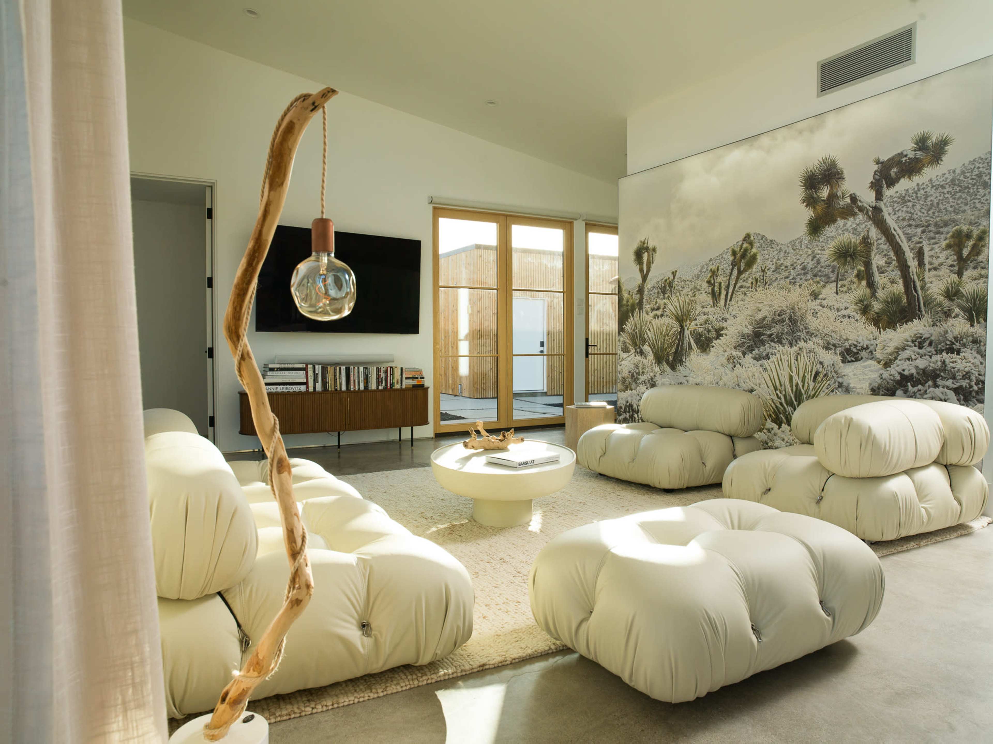 The living room features soft, white seating, a round coffee table, a wall mural of a desert landscape, and large sliding doors leading to an outdoor space.
