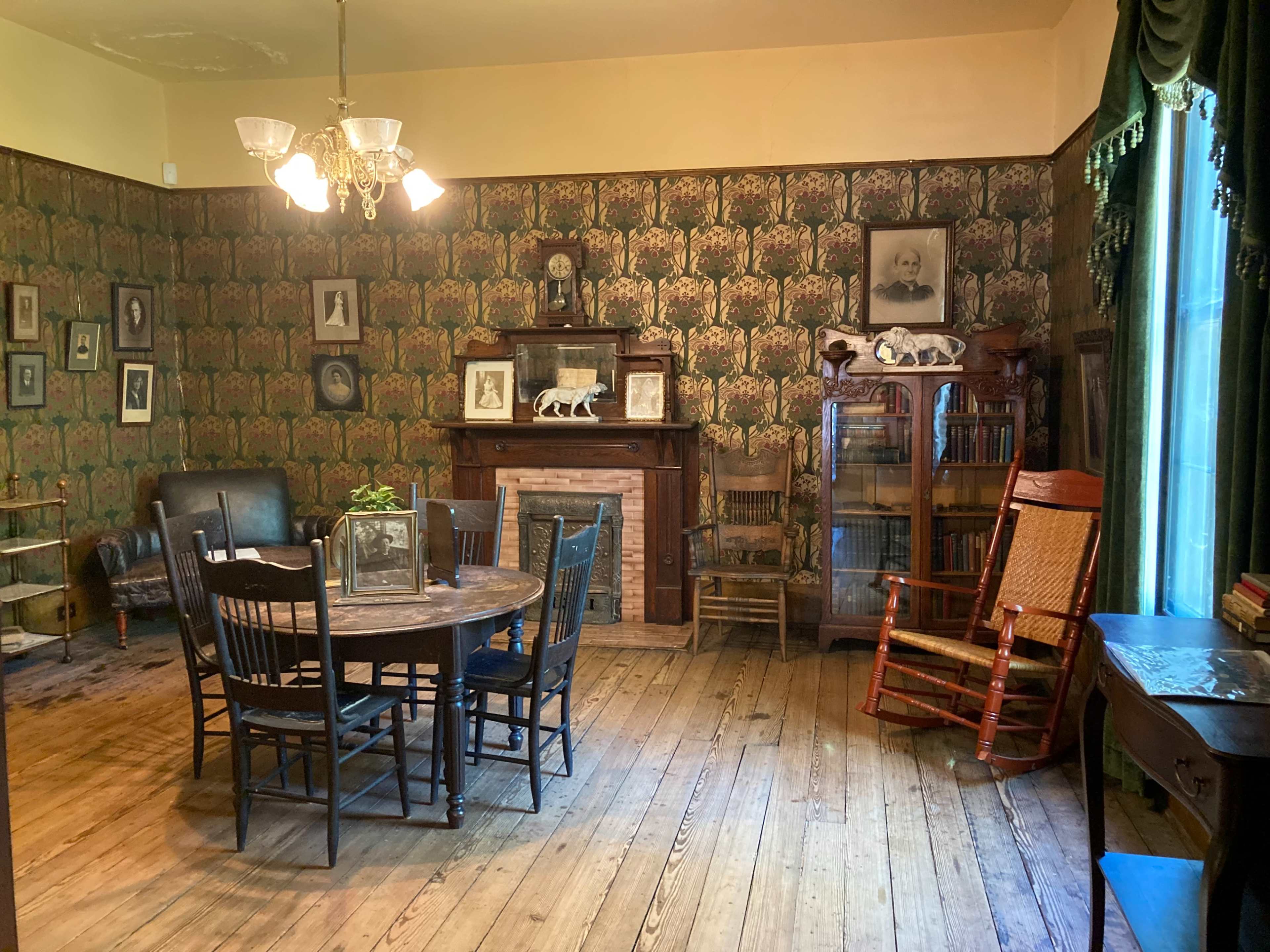 A vintage room with patterned wallpaper featuring a round dining table surrounded by black wooden chairs, a rocking chair, and a fireplace adorned with photographs and decorative items.