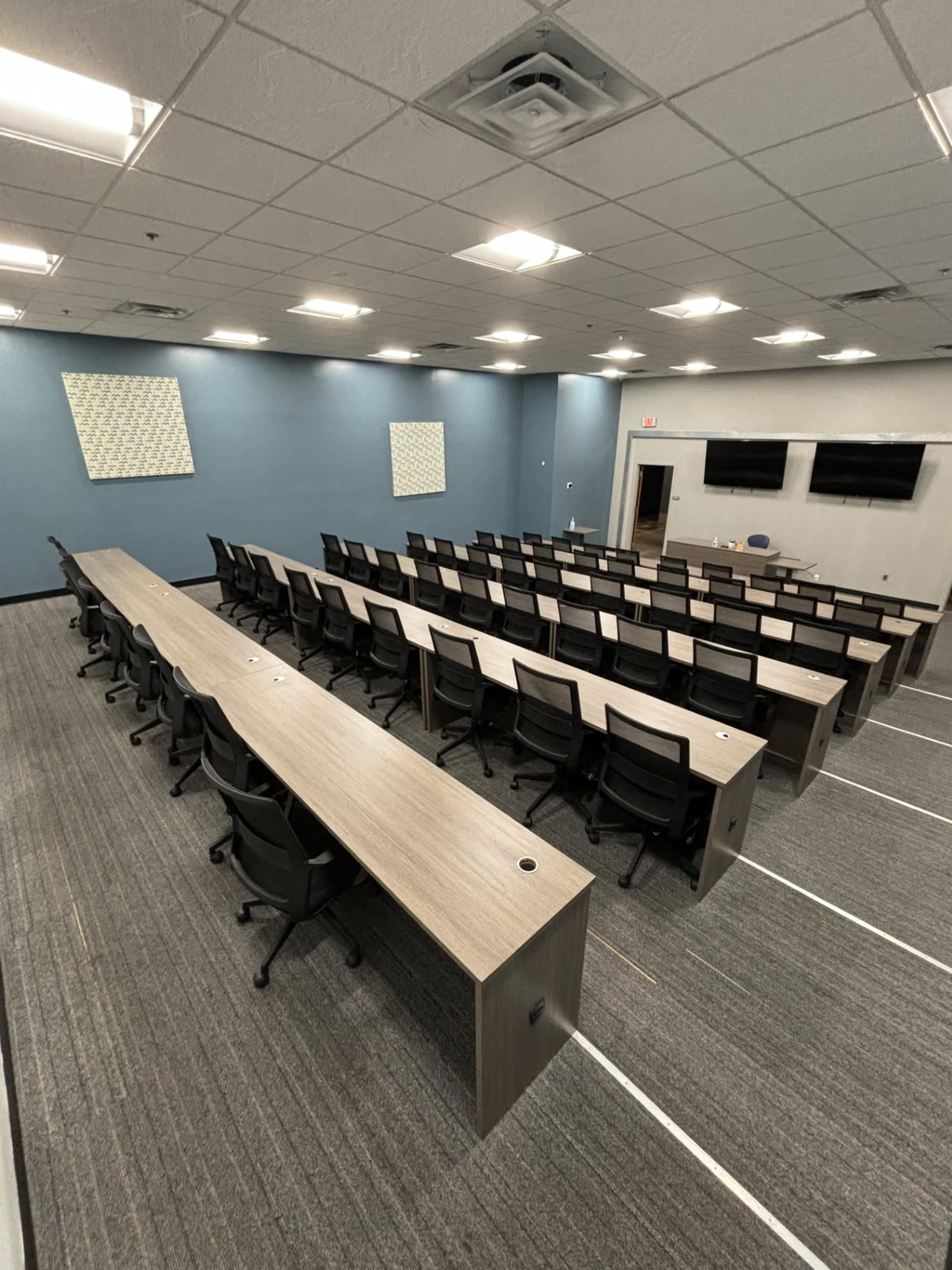 A large conference room arranged with multiple rows of long tables and black chairs facing two screens on the front wall.
