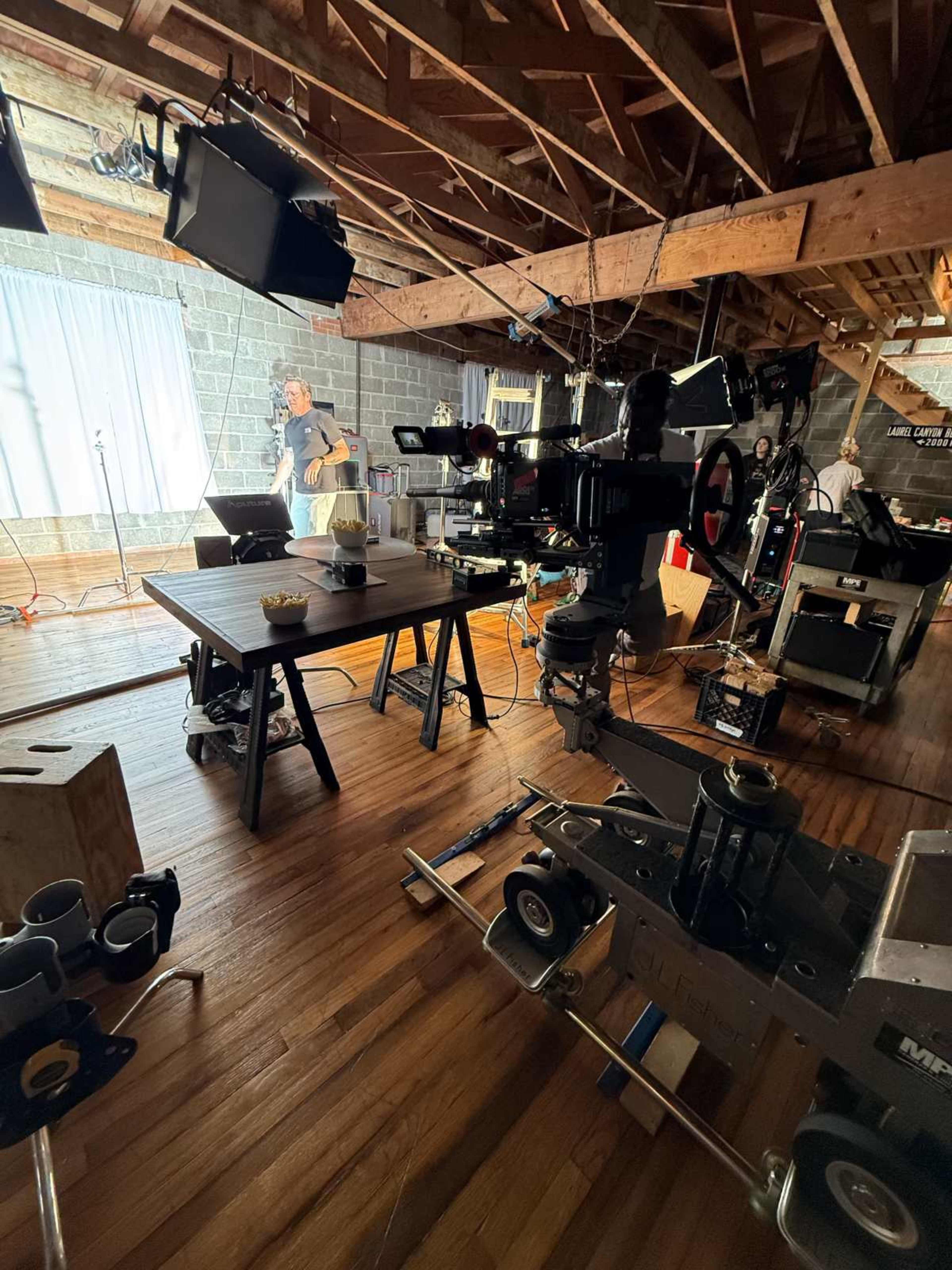 The image shows a partially set-up film studio with various equipment, including cameras and lights, in a rustic interior space.