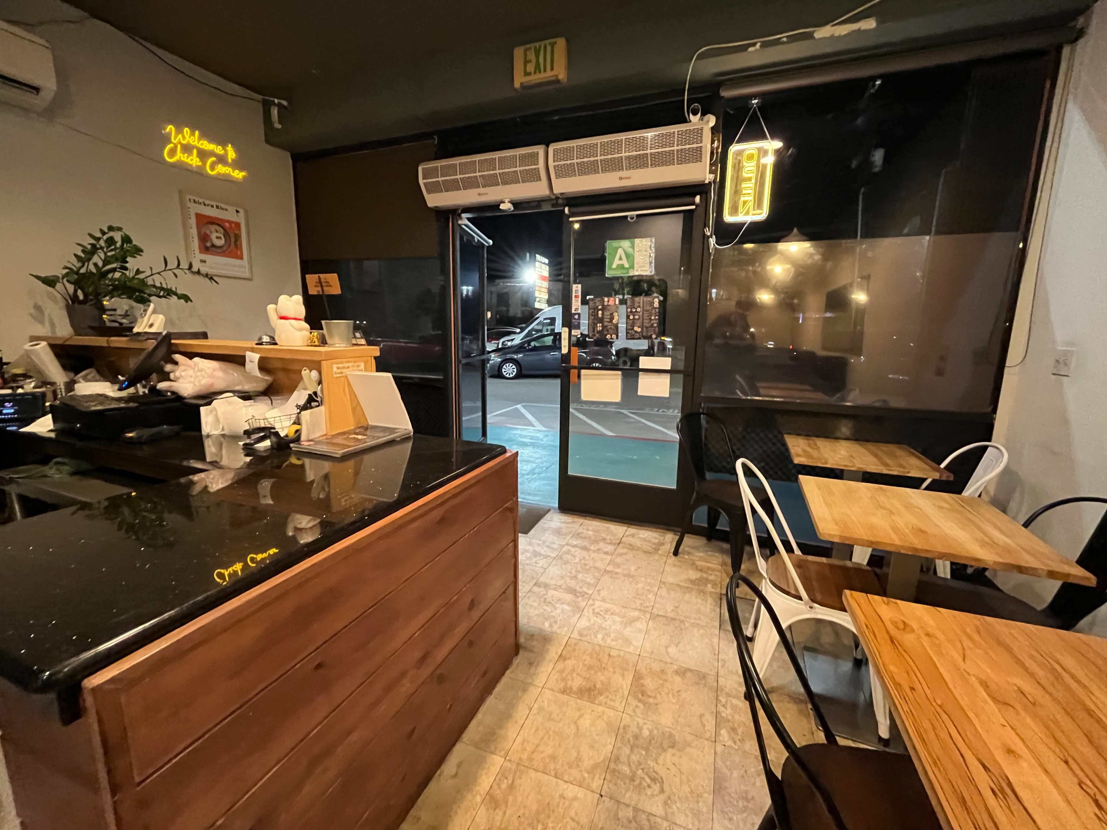 The image shows the interior of a small café with wooden furniture, a counter with a cash register, and a view of the entrance door illuminated by an open sign.