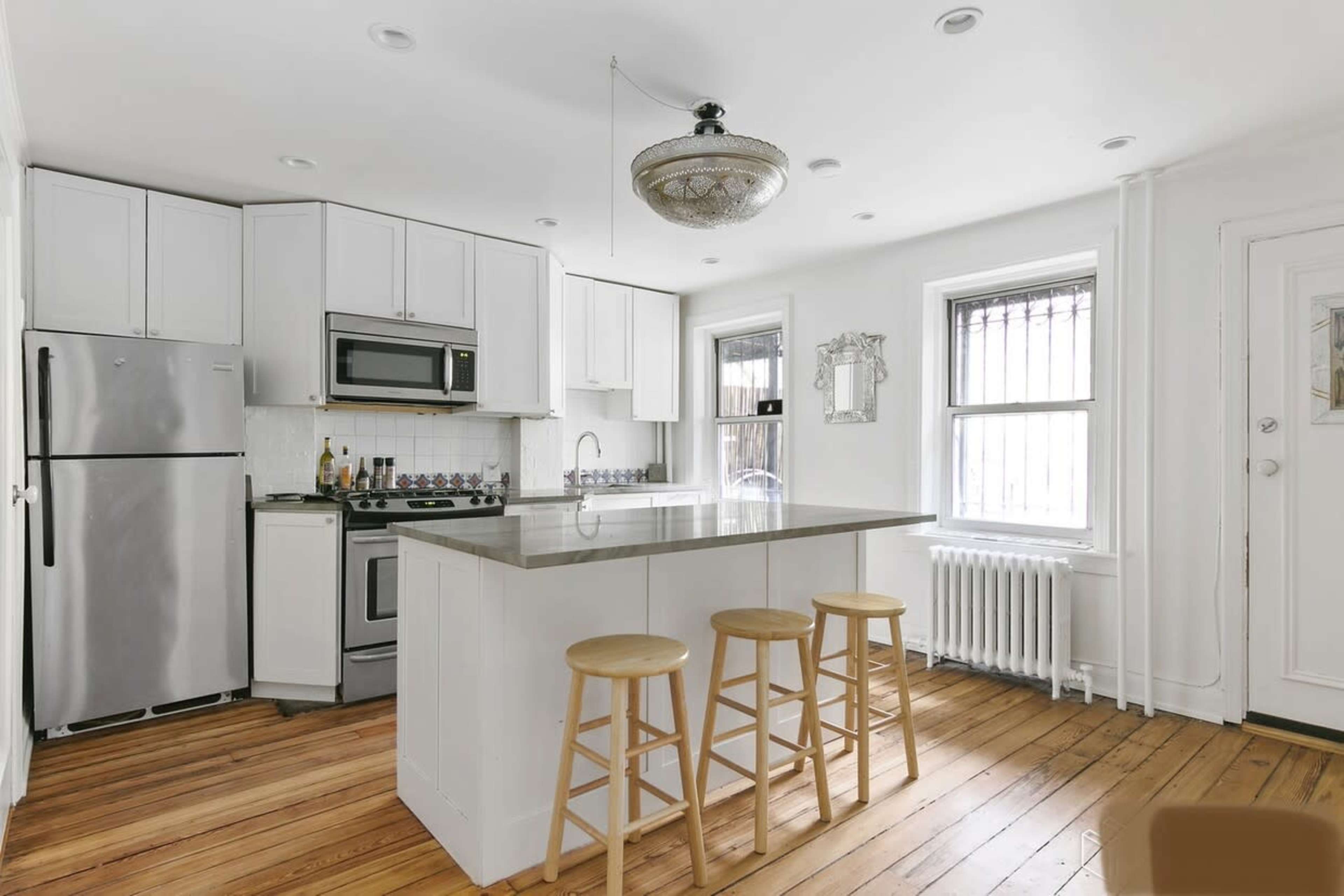 A modern kitchen with white cabinetry, stainless steel appliances, a central island with three wooden stools, and hardwood flooring.