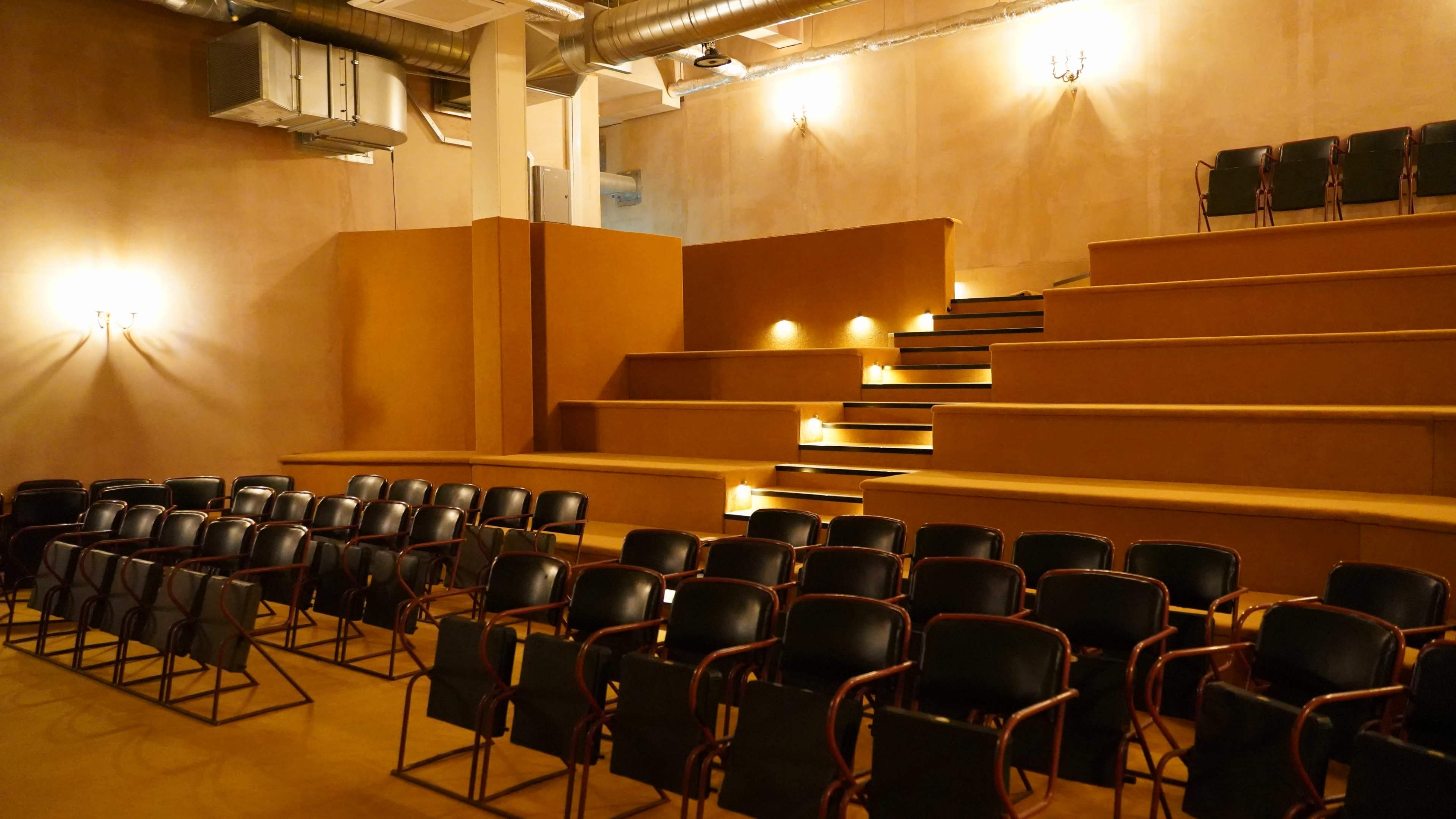 The image shows a semi-modern theater with tiered seating and black chairs arranged in rows facing a stage area illuminated by soft lighting.