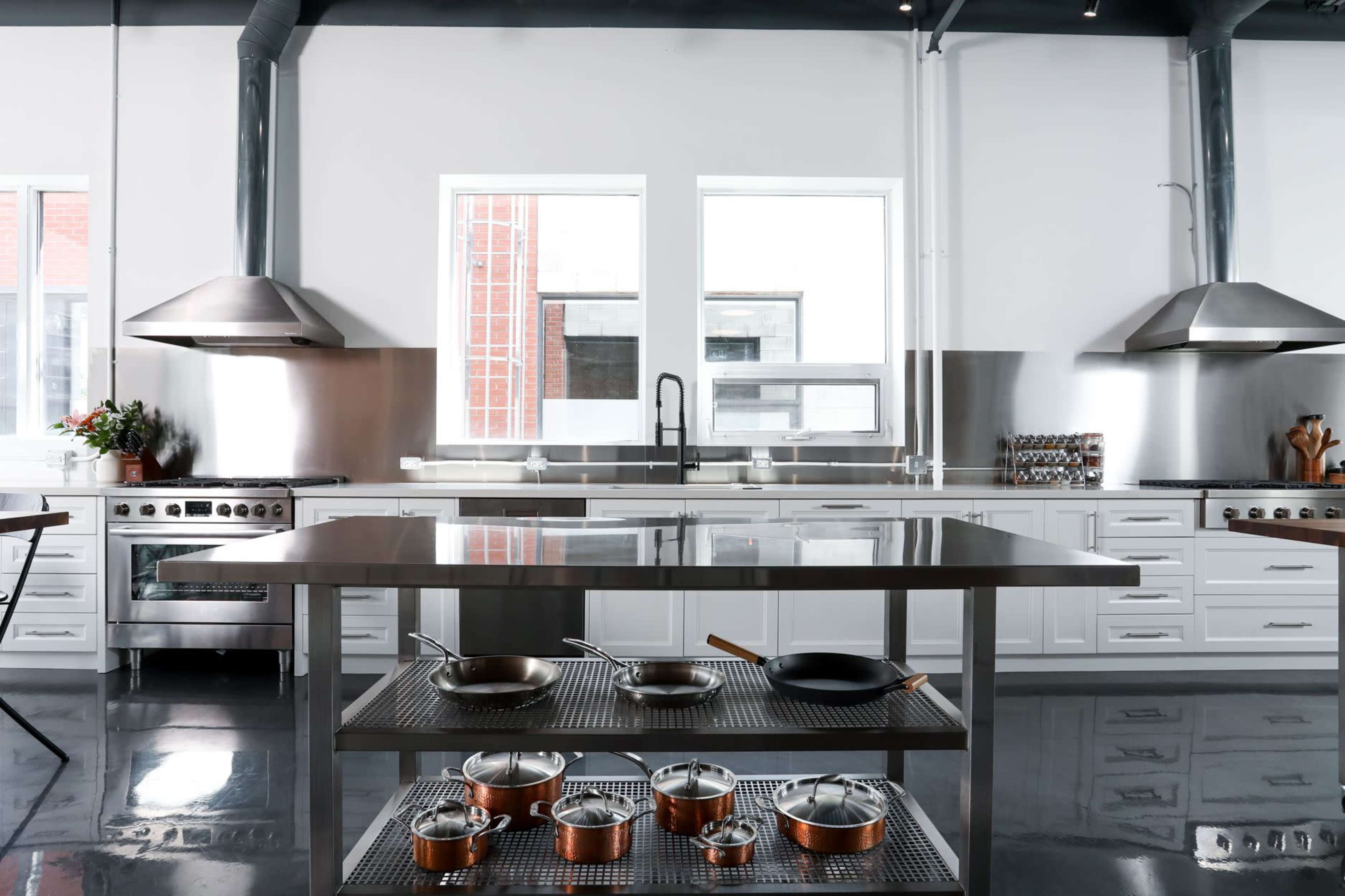 The image shows a modern kitchen with stainless steel appliances, a central island with a metal frame, and various cooking pots arranged on open shelves.