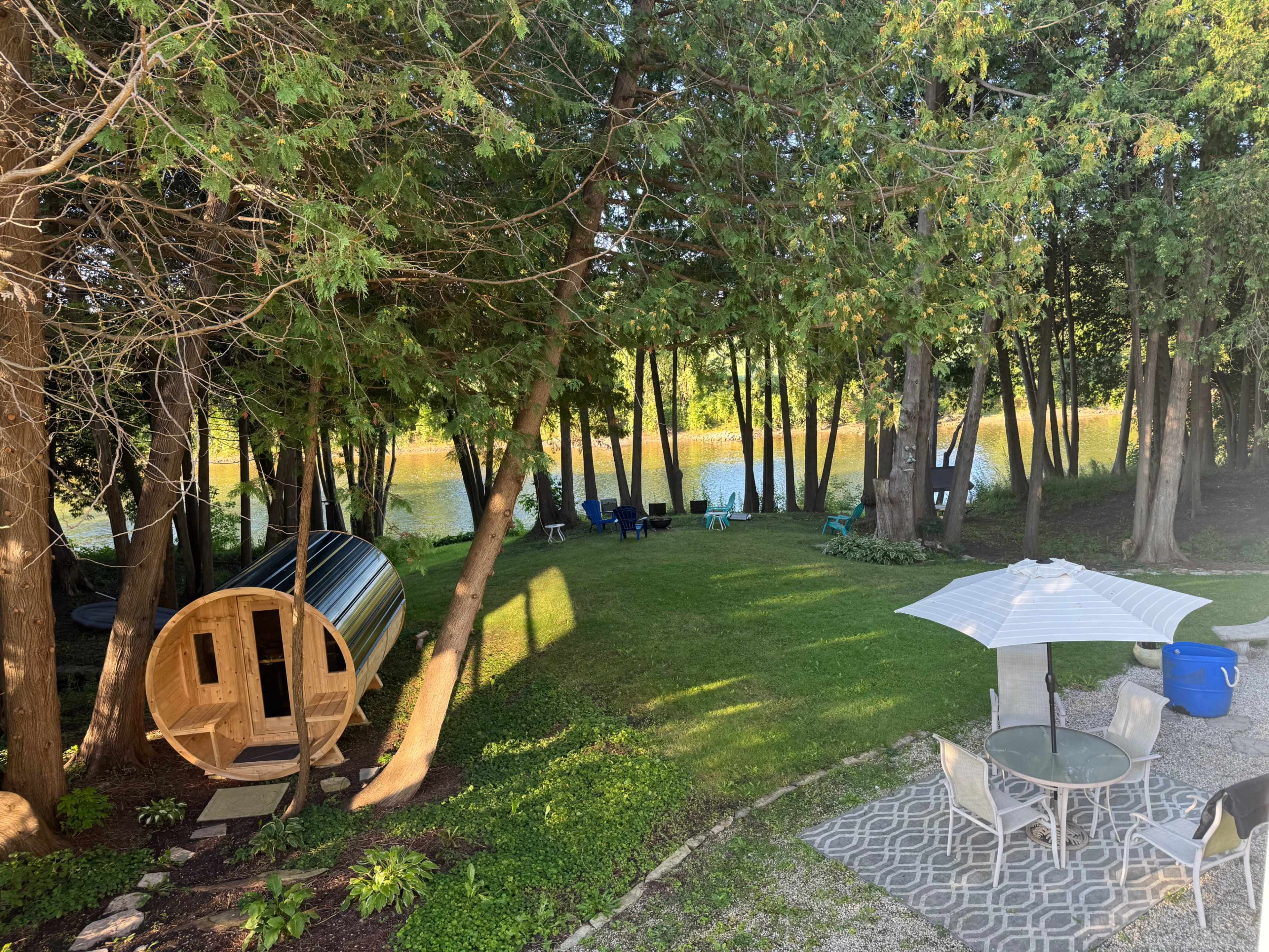 The scene shows a backyard with a wooden sauna, a patio set under an umbrella, and a view of a calm river flanked by trees.