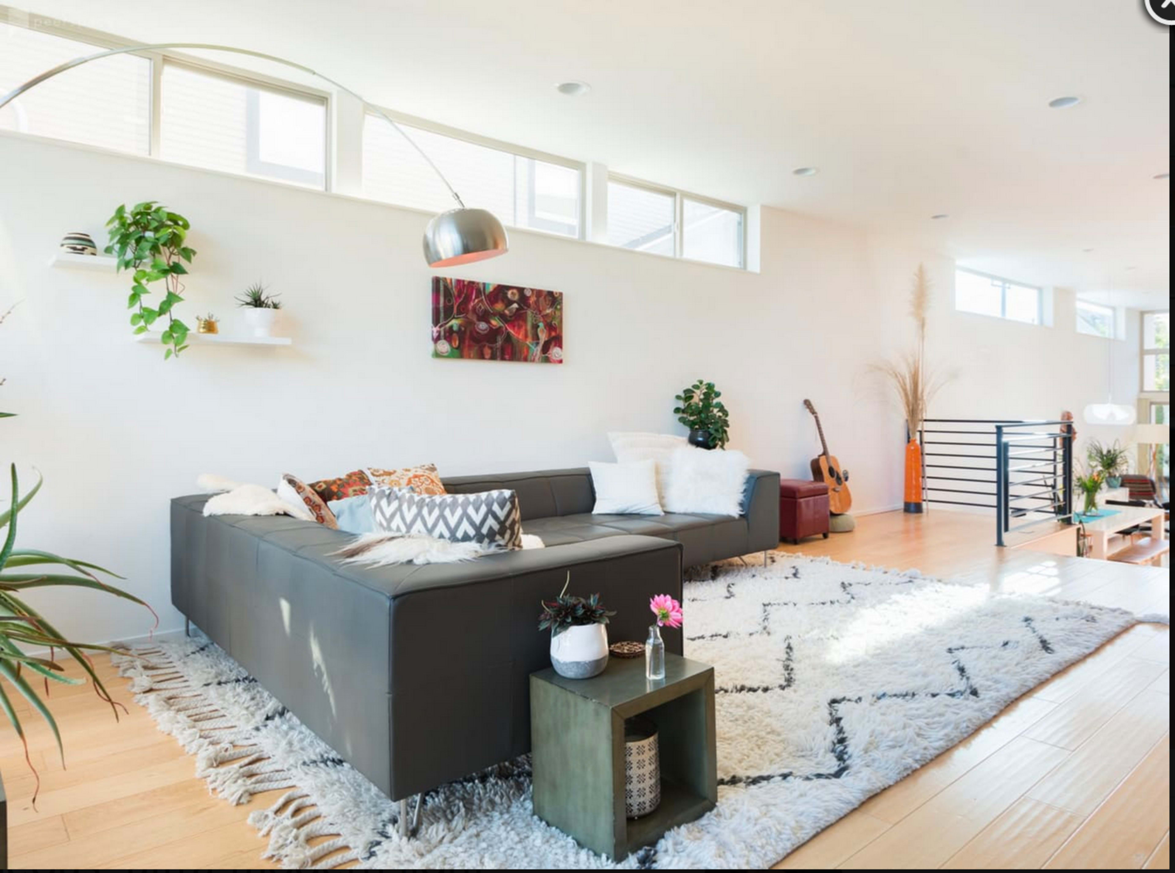 The living room features a sectional sofa, decorative plants, and a coffee table on a large, textured rug, with guitar and various decorative elements in the corner.