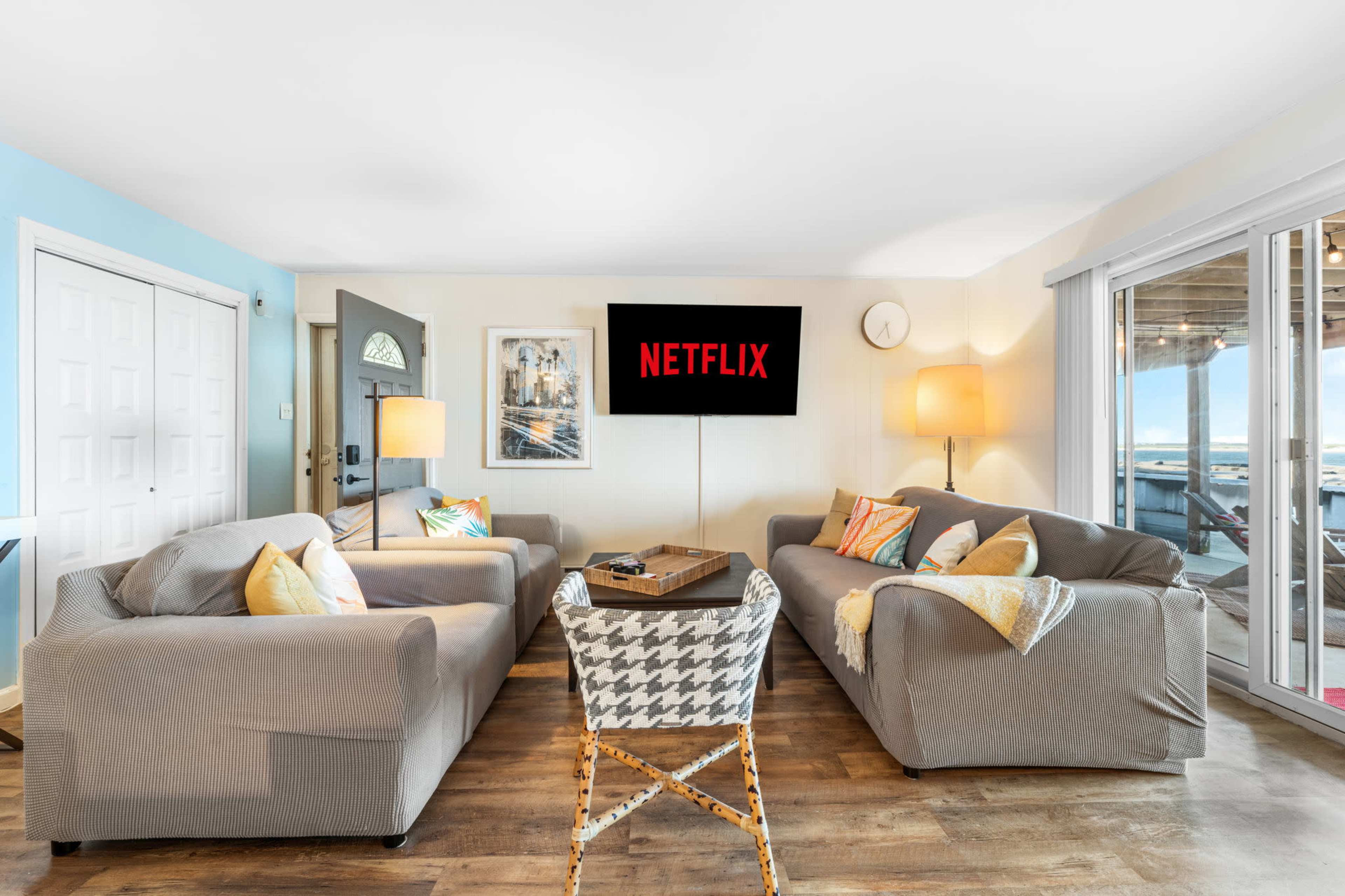 A living room features two gray sofas, a coffee table, a wall-mounted television displaying the Netflix logo, and large sliding glass doors leading to an outdoor area.