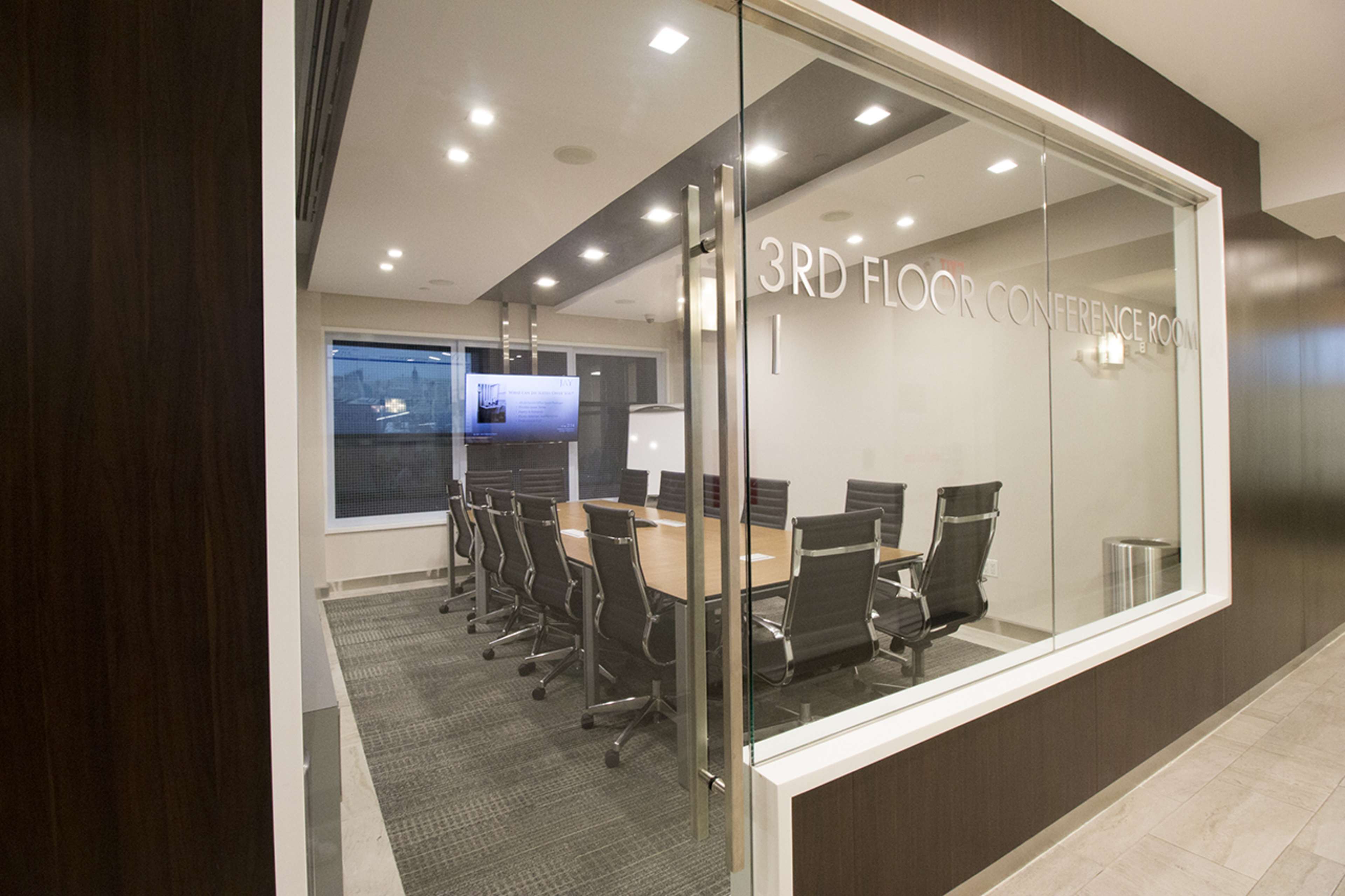 A modern conference room with a long table surrounded by black chairs, visible through glass walls.