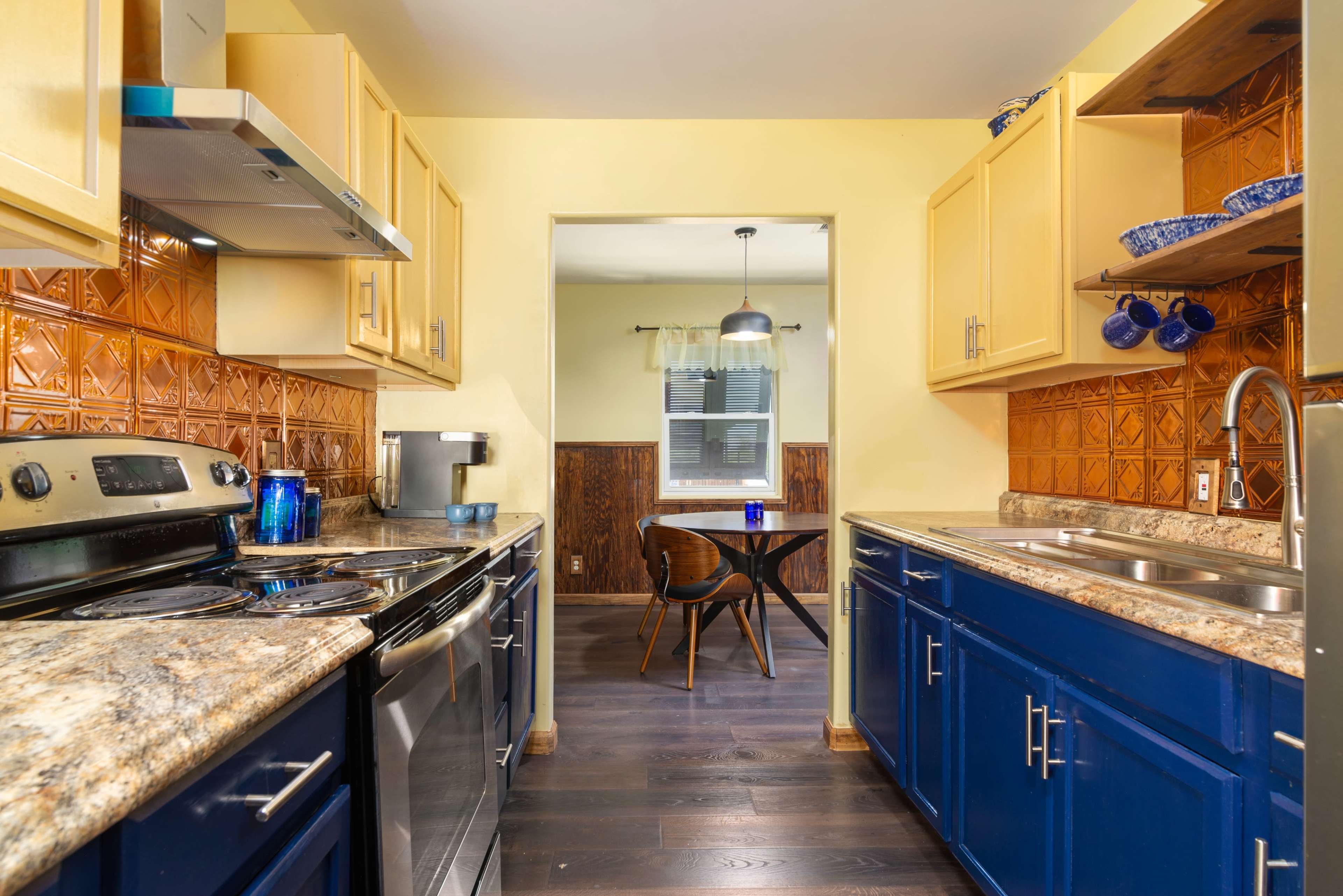 The image shows a compact kitchen with yellow walls, blue cabinets, and granite countertops, leading to a dining area through an archway.