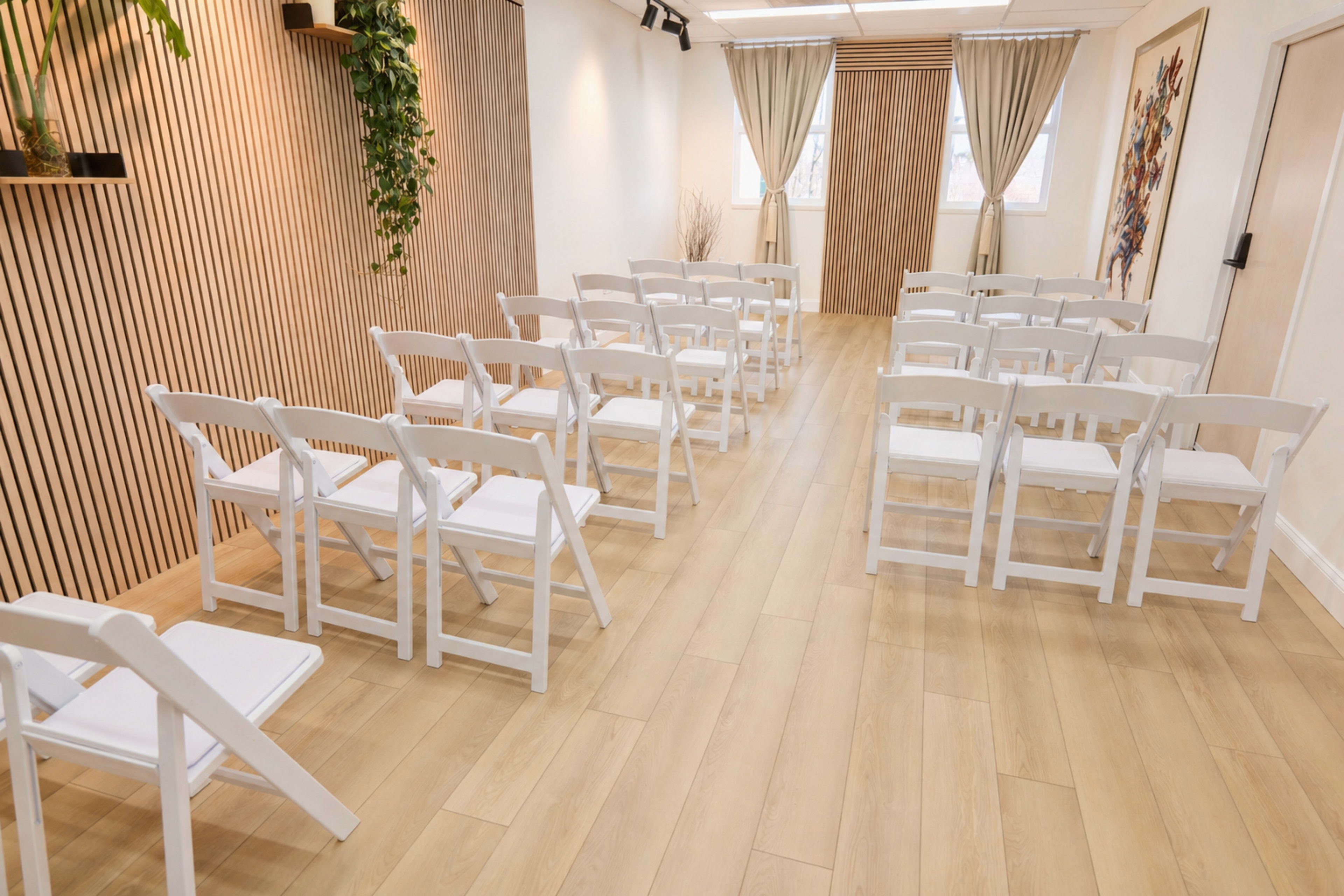 The image shows a bright room set up with white folding chairs arranged in rows facing a wooden accent wall.