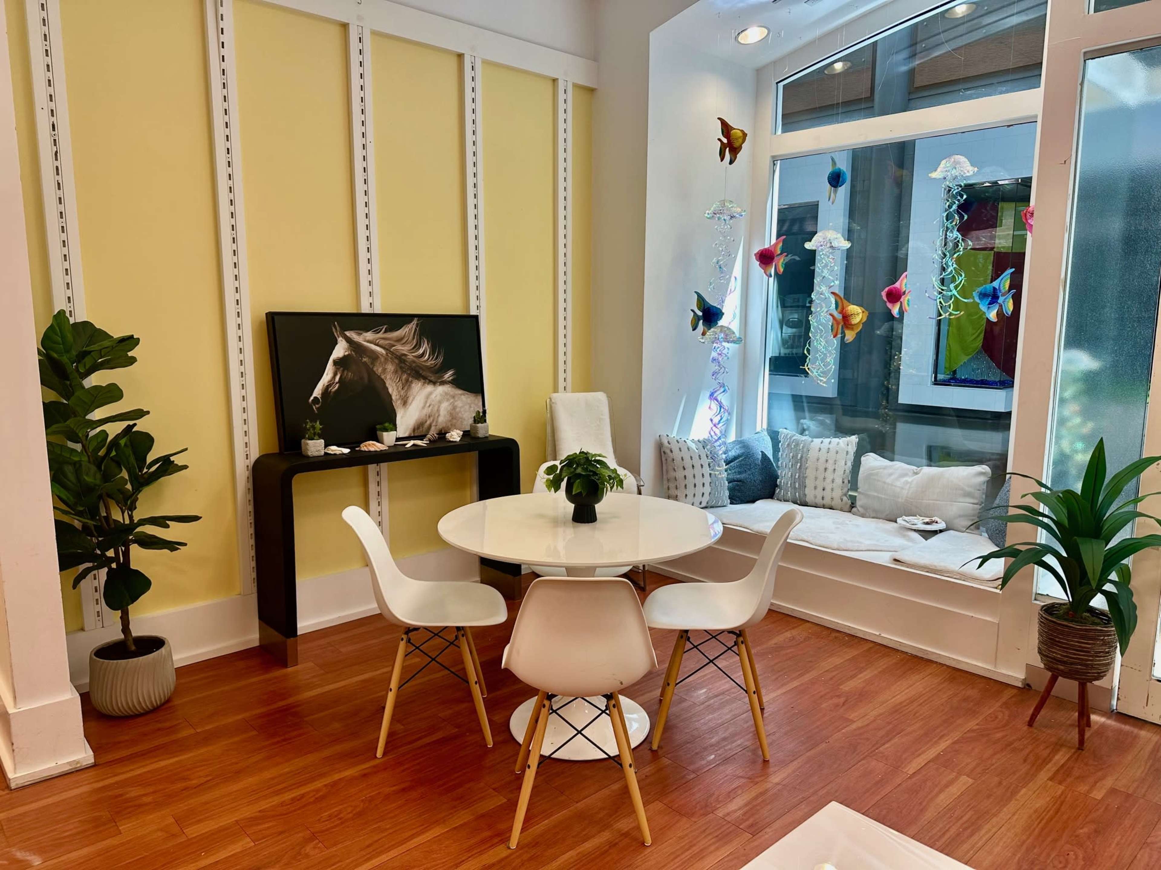 The image shows a bright indoor space featuring a circular white dining table with four chairs, a black console table displaying a horse photograph, and a cozy window nook adorned with decorative butterflies and jellyfish designs.