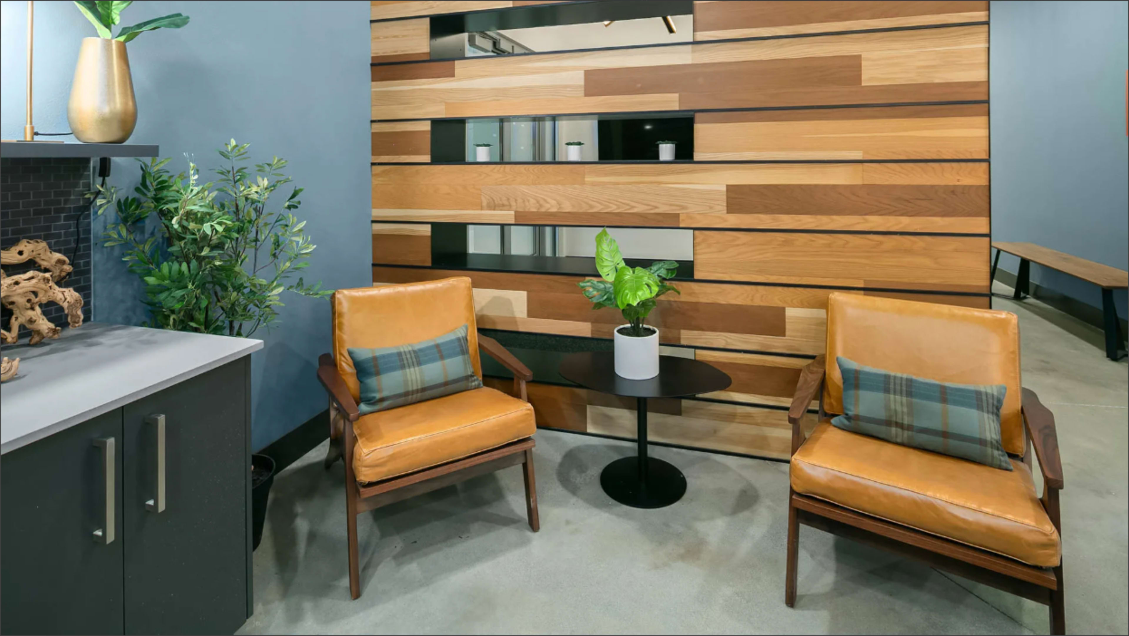 The image shows a modern sitting area with two leather armchairs, a small round table, and a wooden accent wall, complemented by a potted plant.