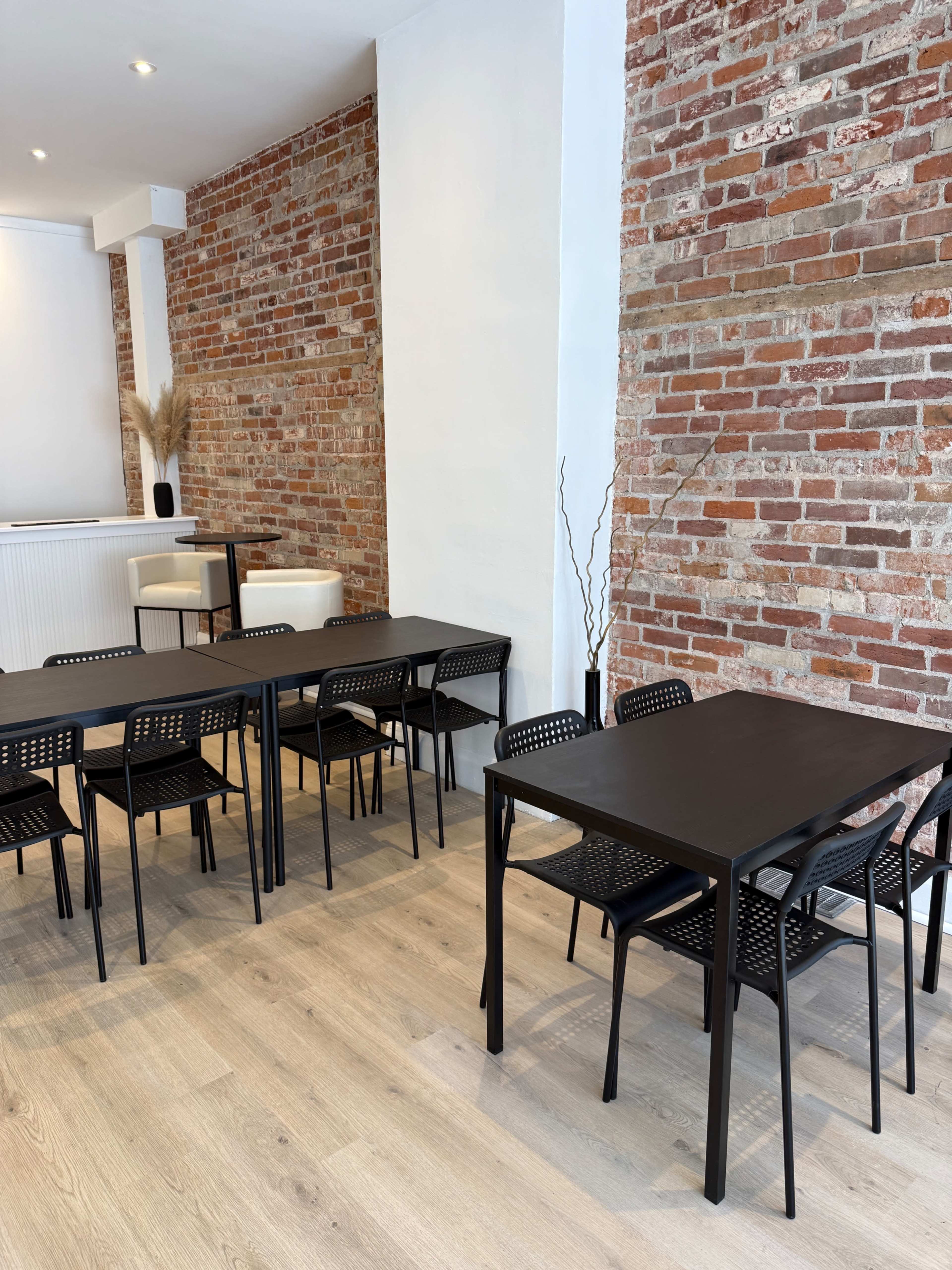 The image shows a modern interior space with exposed brick walls, featuring black tables and chairs arranged neatly on a light wooden floor.