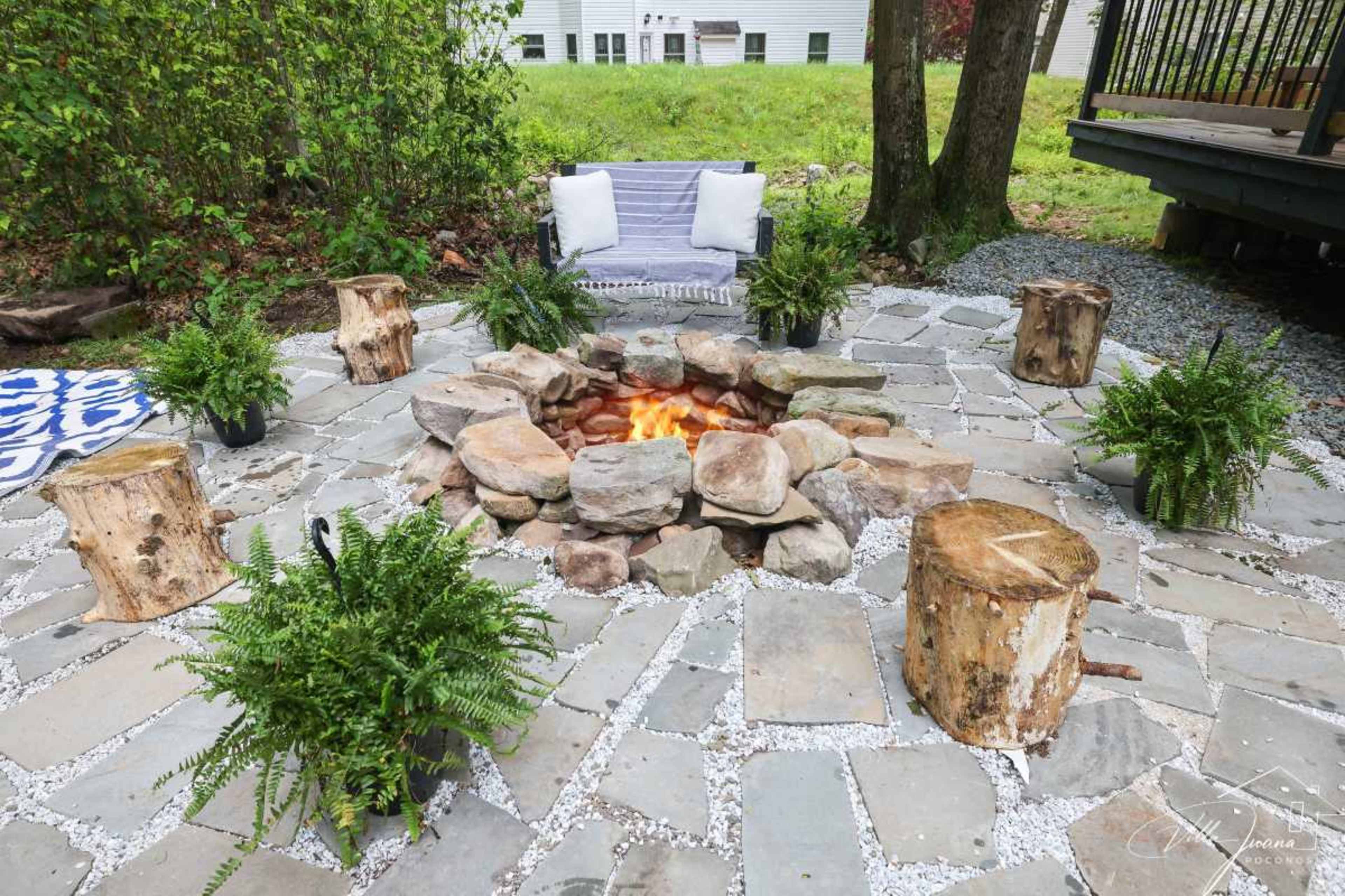 The image shows a circular stone fire pit surrounded by wooden stumps as seats and potted ferns, set in a grassy outdoor area.