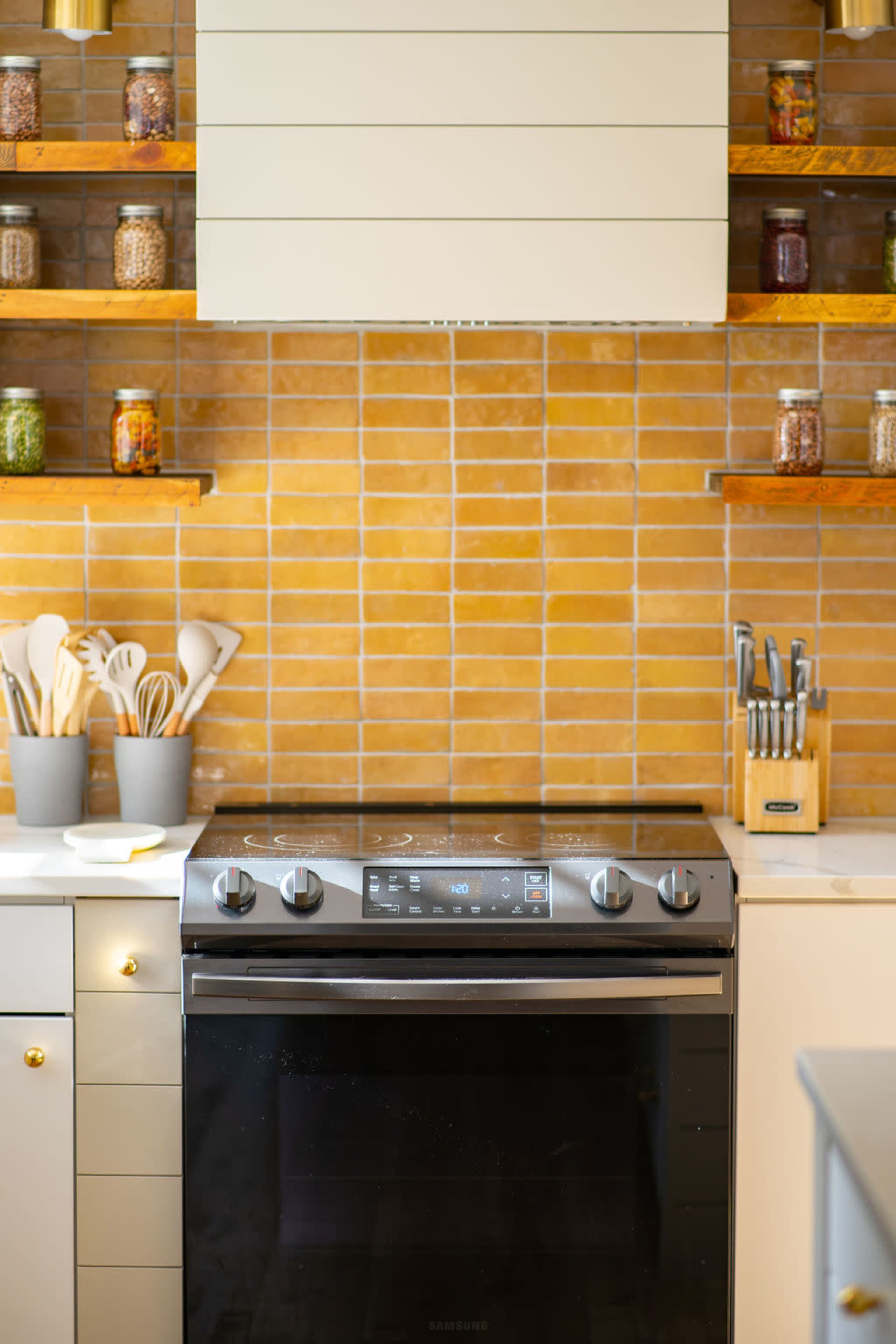 A modern kitchen featuring a black stove beneath a hood, with wooden shelves displaying jars and utensils above a light-colored countertop.