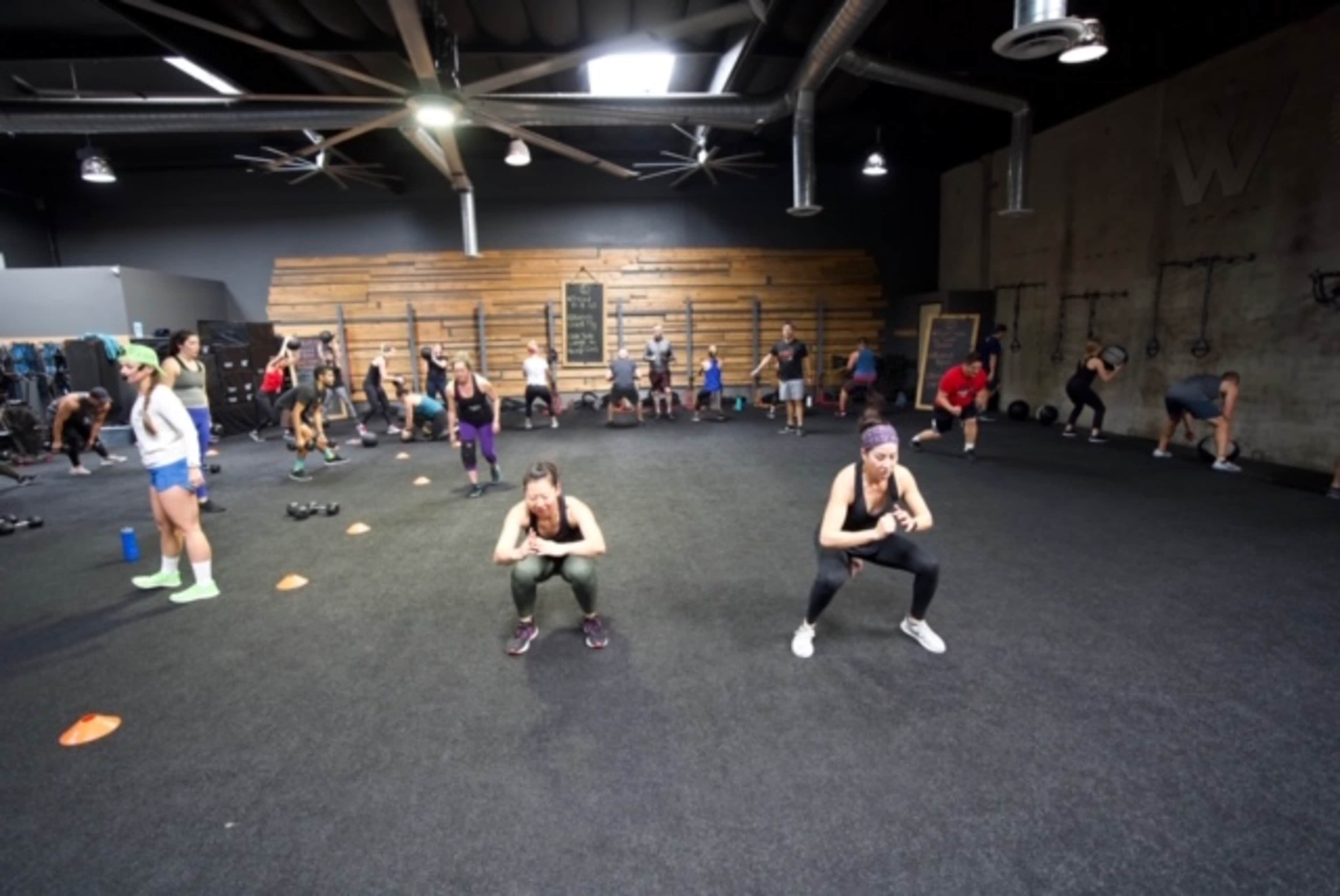 A group of people is participating in a fitness class at a gym, with some performing squats on the black floor while others engage in various exercises around them.