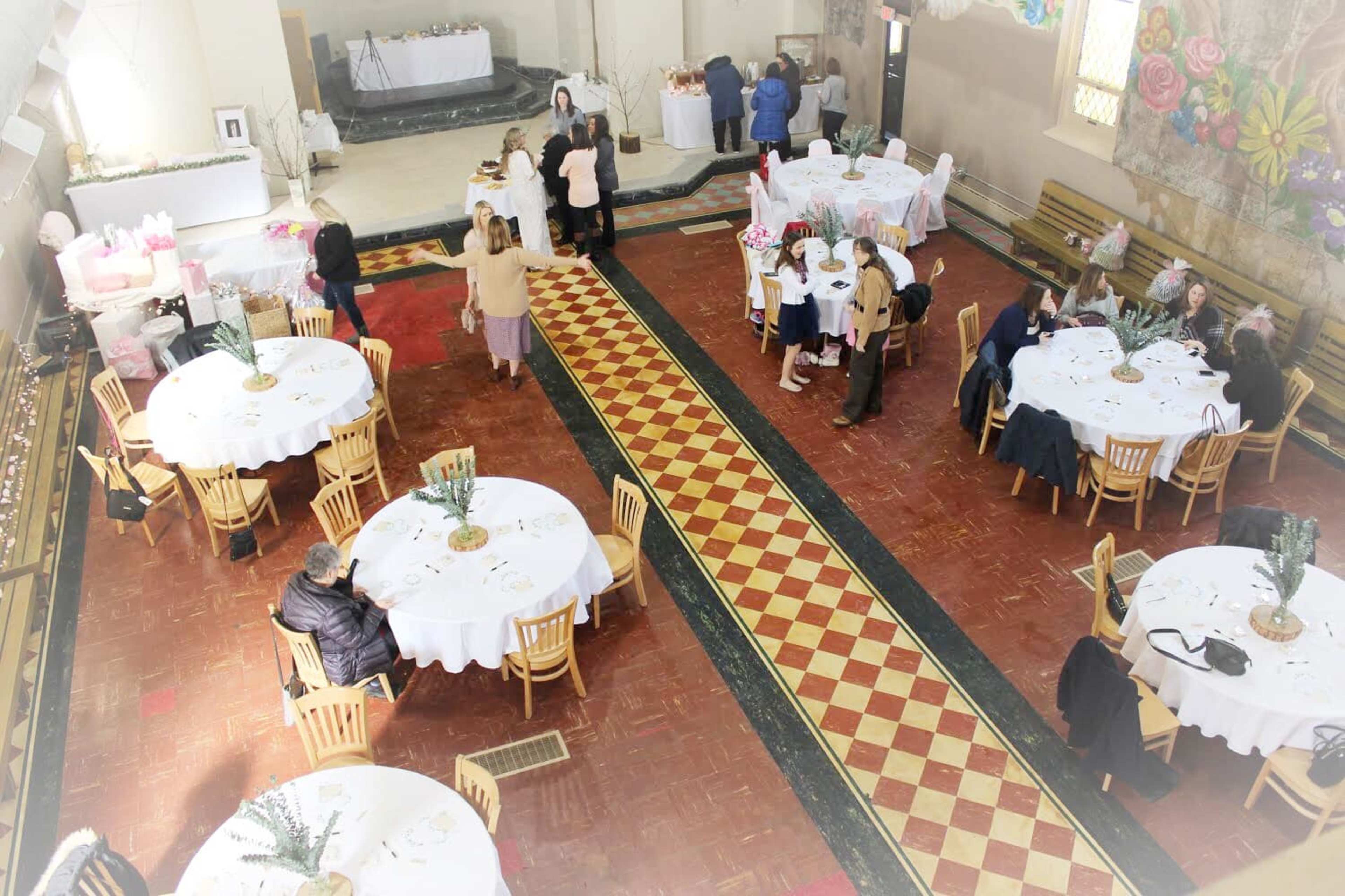 A spacious event hall is set up with round tables covered in white tablecloths, with guests mingling and a buffet area in the background.