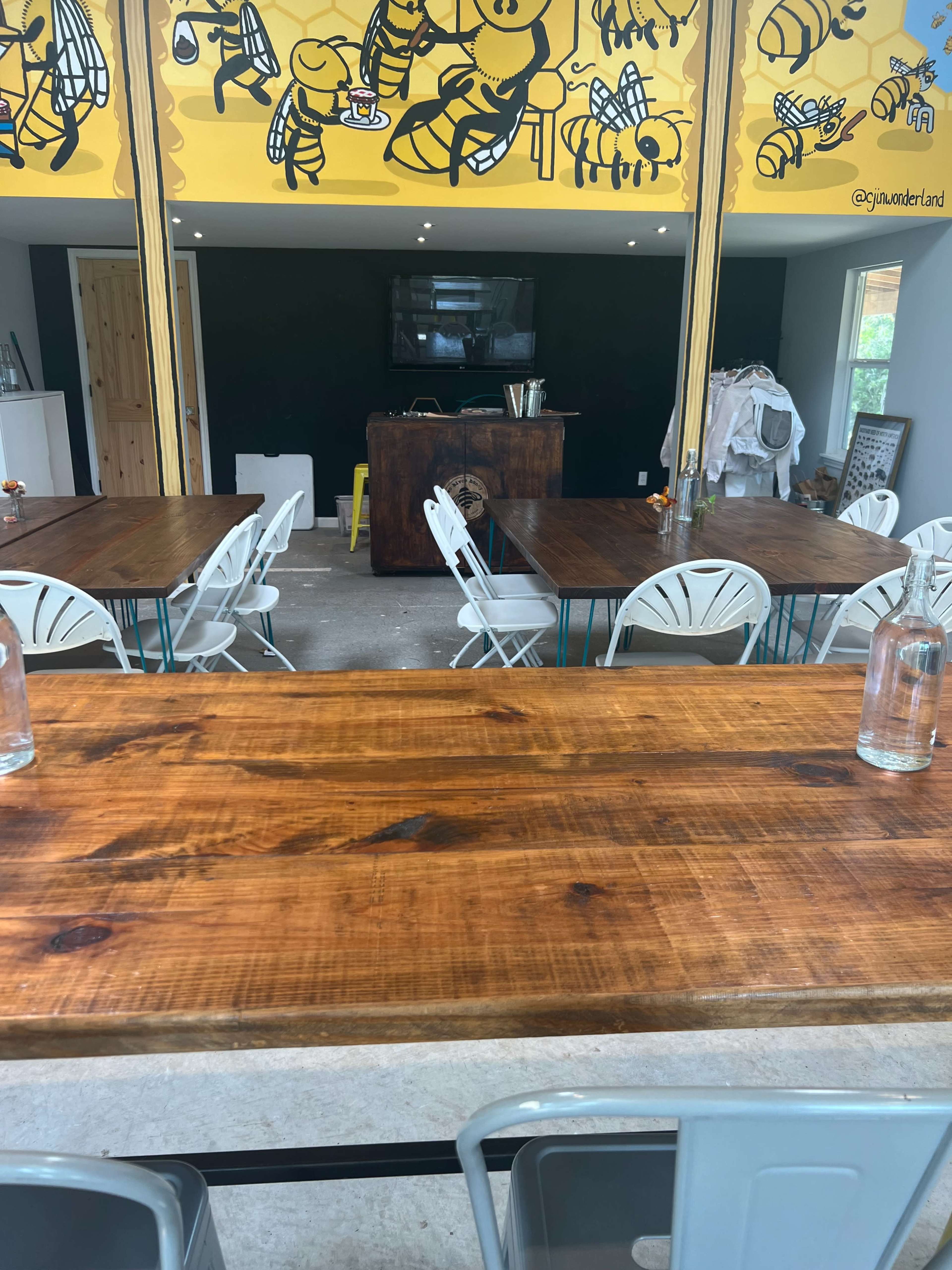 The image shows a room with wooden tables and white chairs, featuring a large mural of bees on the wall and a beverage station at the back.