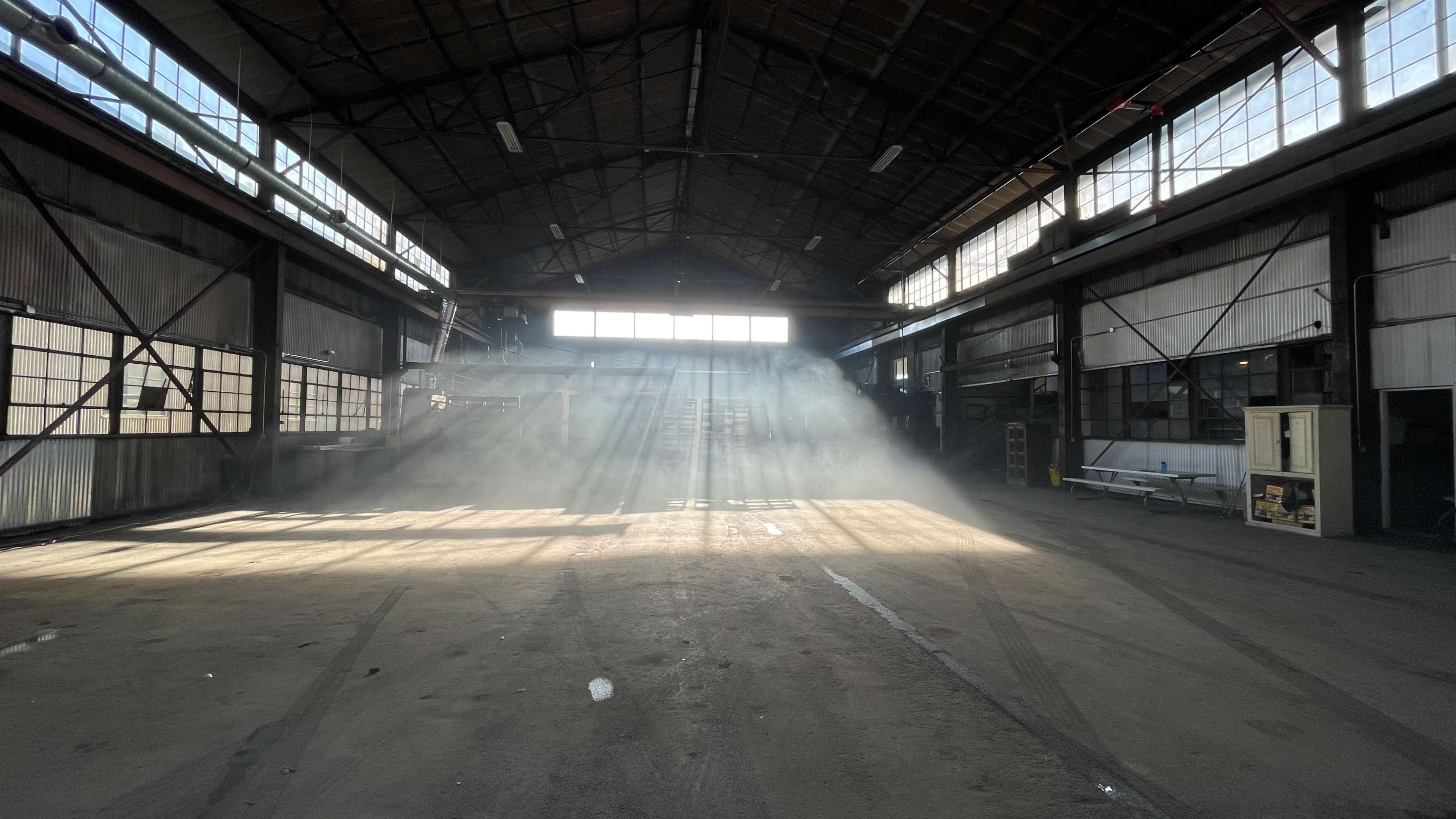 Sunlight streams through the windows of a large, empty warehouse, creating beams of light in the dust-filled air.