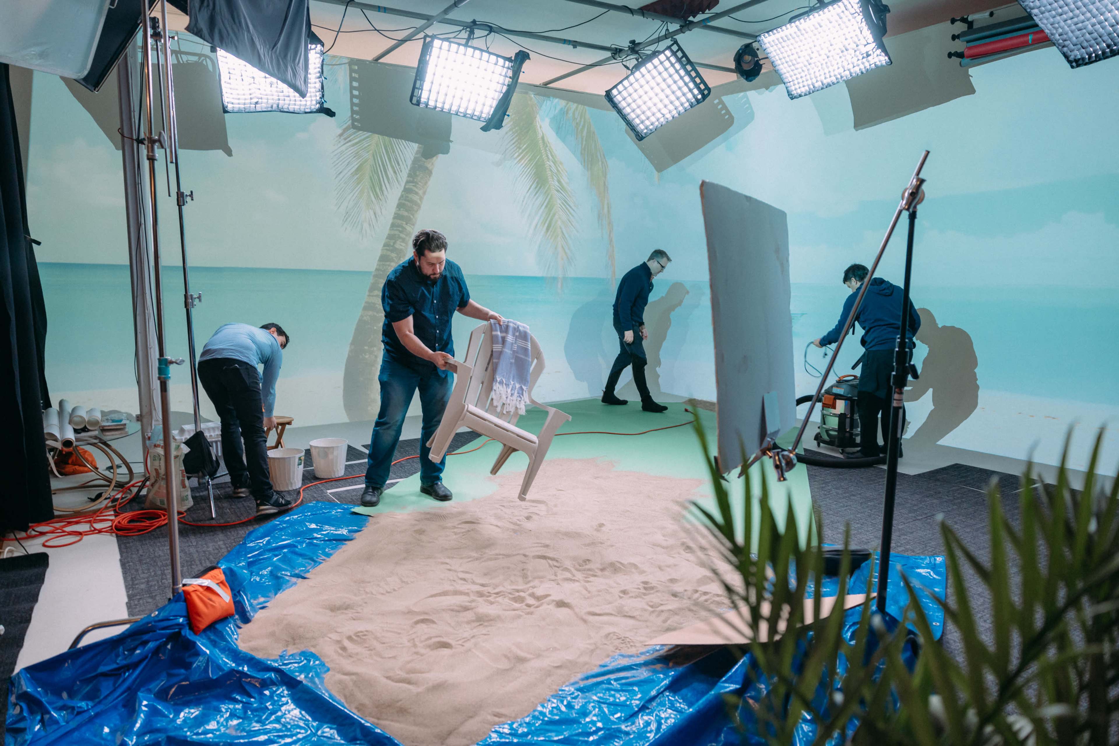 Two crew members set up a tropical-themed film set with a sandy area and backdrop, while another crew member adjusts equipment.