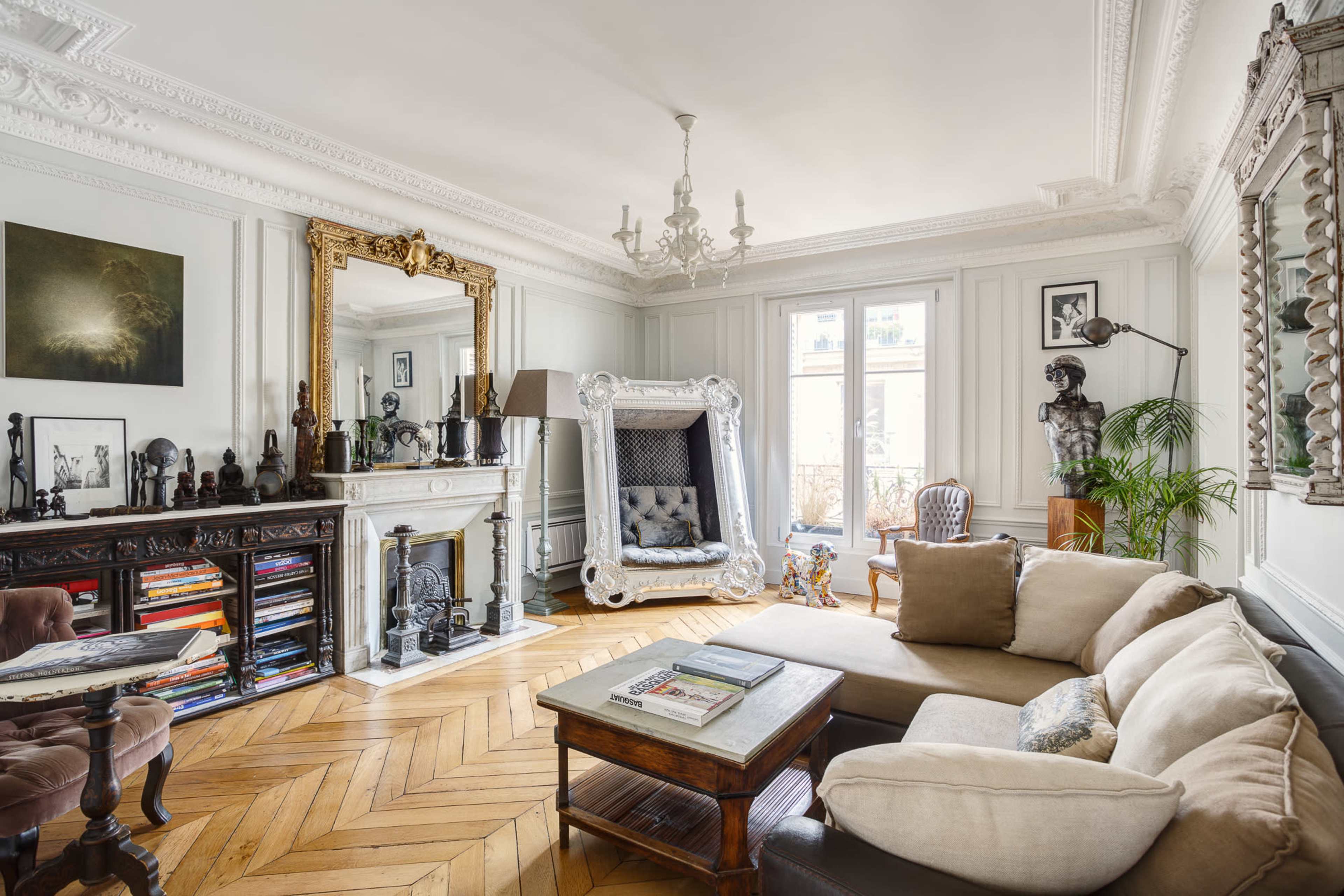 A stylish living room with a white couch, hardwood flooring, ornate molding, and decorative furnishings including a large mirror and various sculptures.
