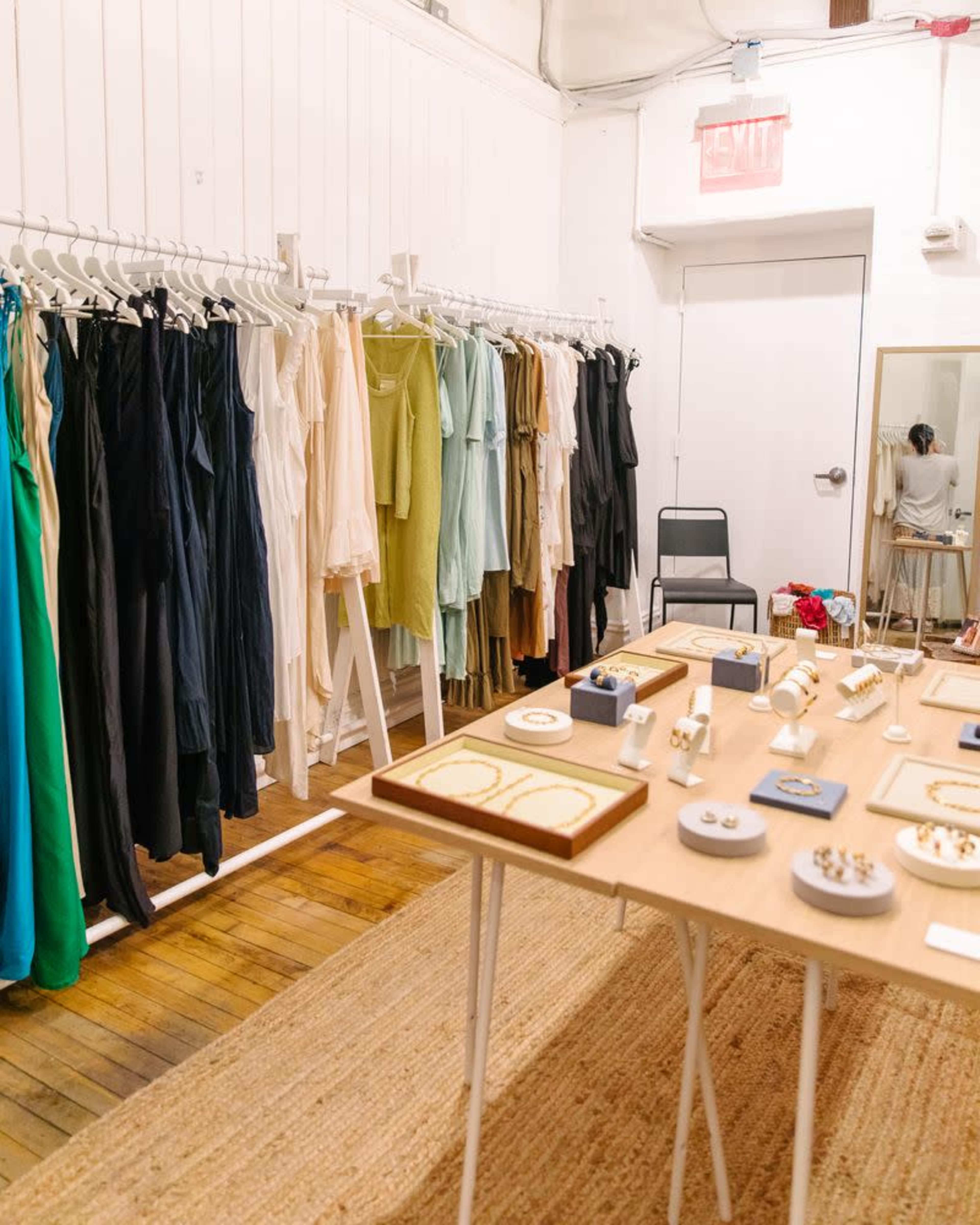 The image shows a clothing and jewelry display in a bright room, featuring a rack of dresses in various colors and a table with neatly organized jewelry pieces.