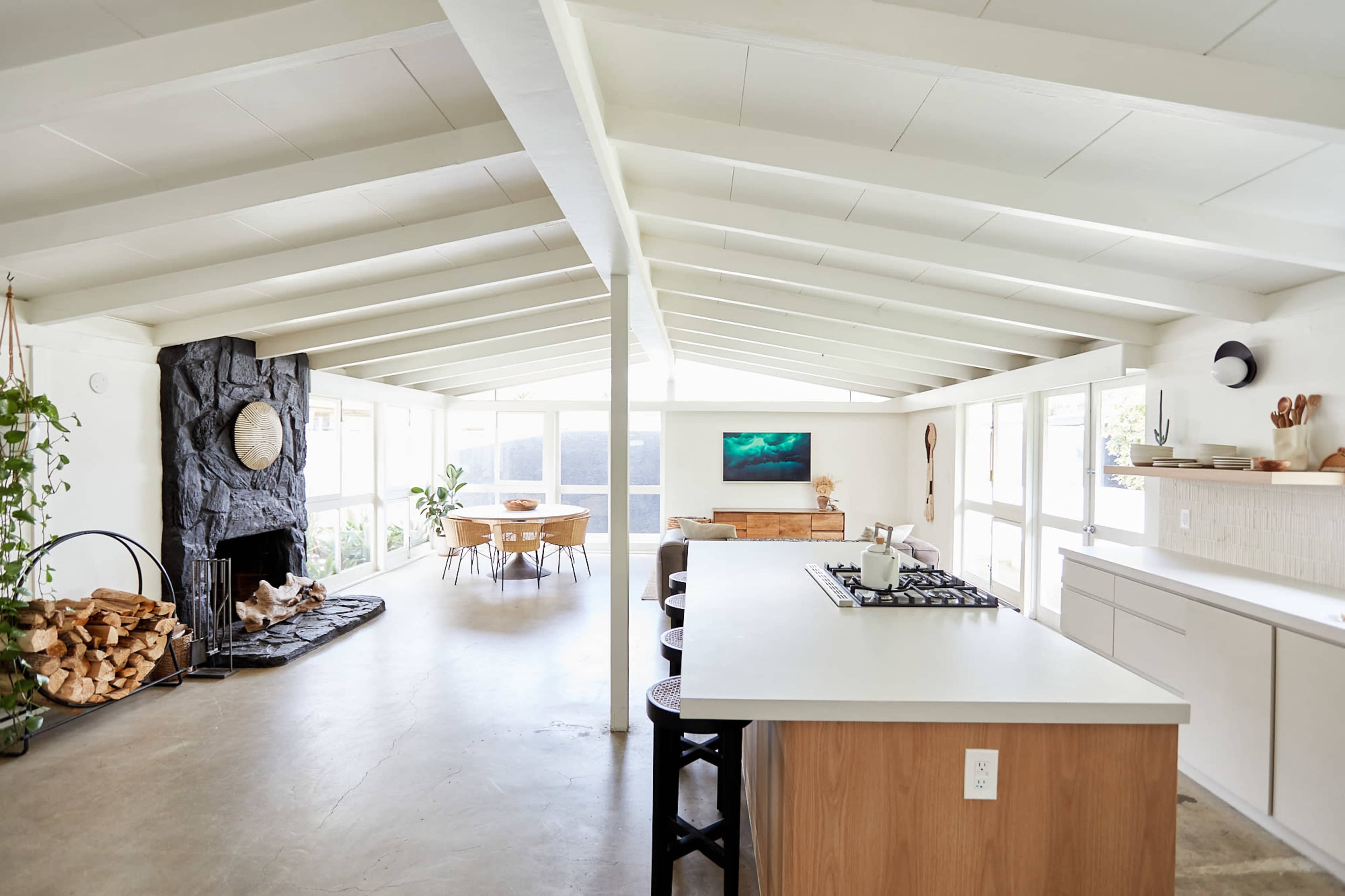 The image shows a modern, open-concept kitchen and living area with a stone fireplace, large windows, and a dining table.