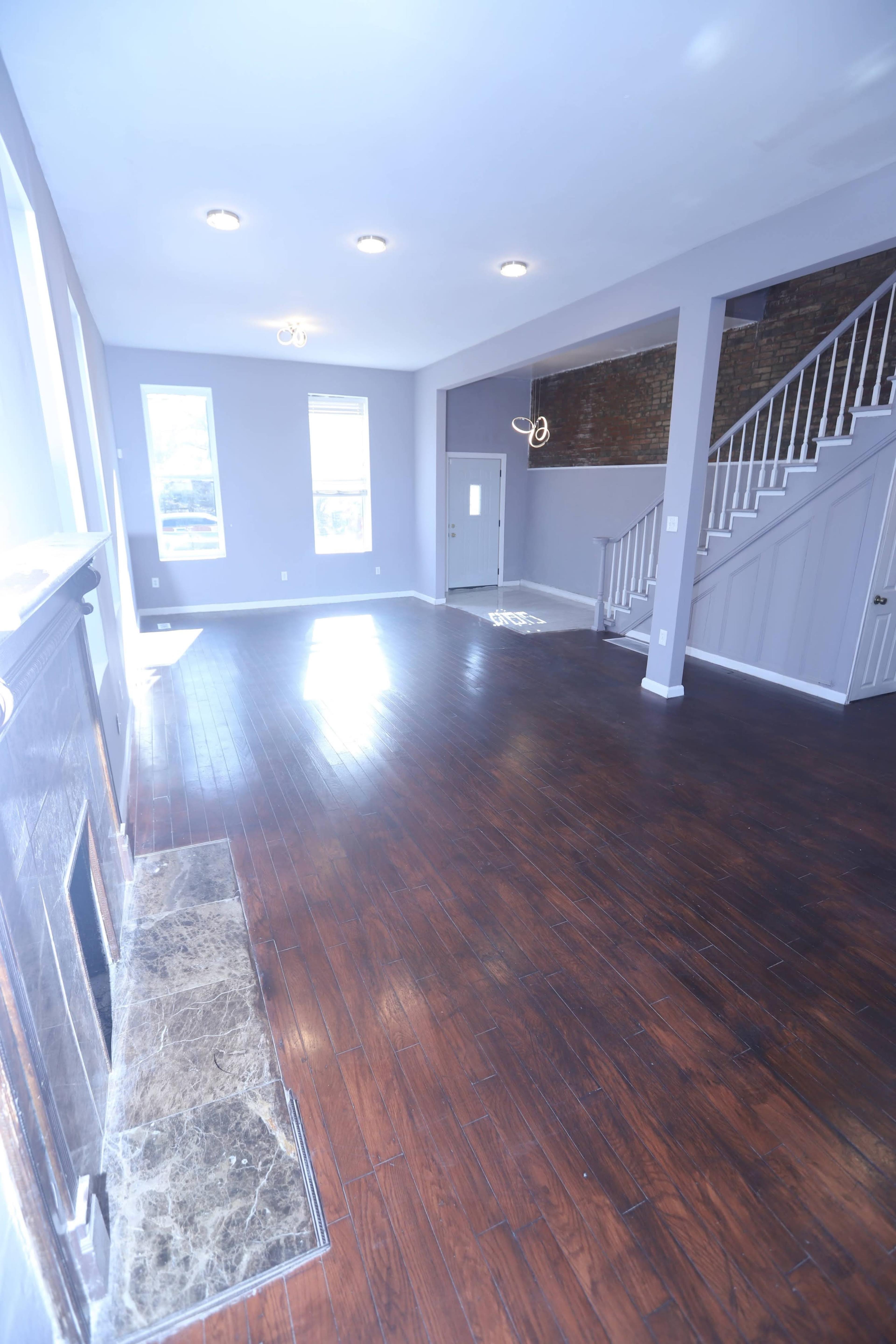 The image shows a spacious living room with wood flooring and a staircase, featuring large windows that allow natural light to enter.