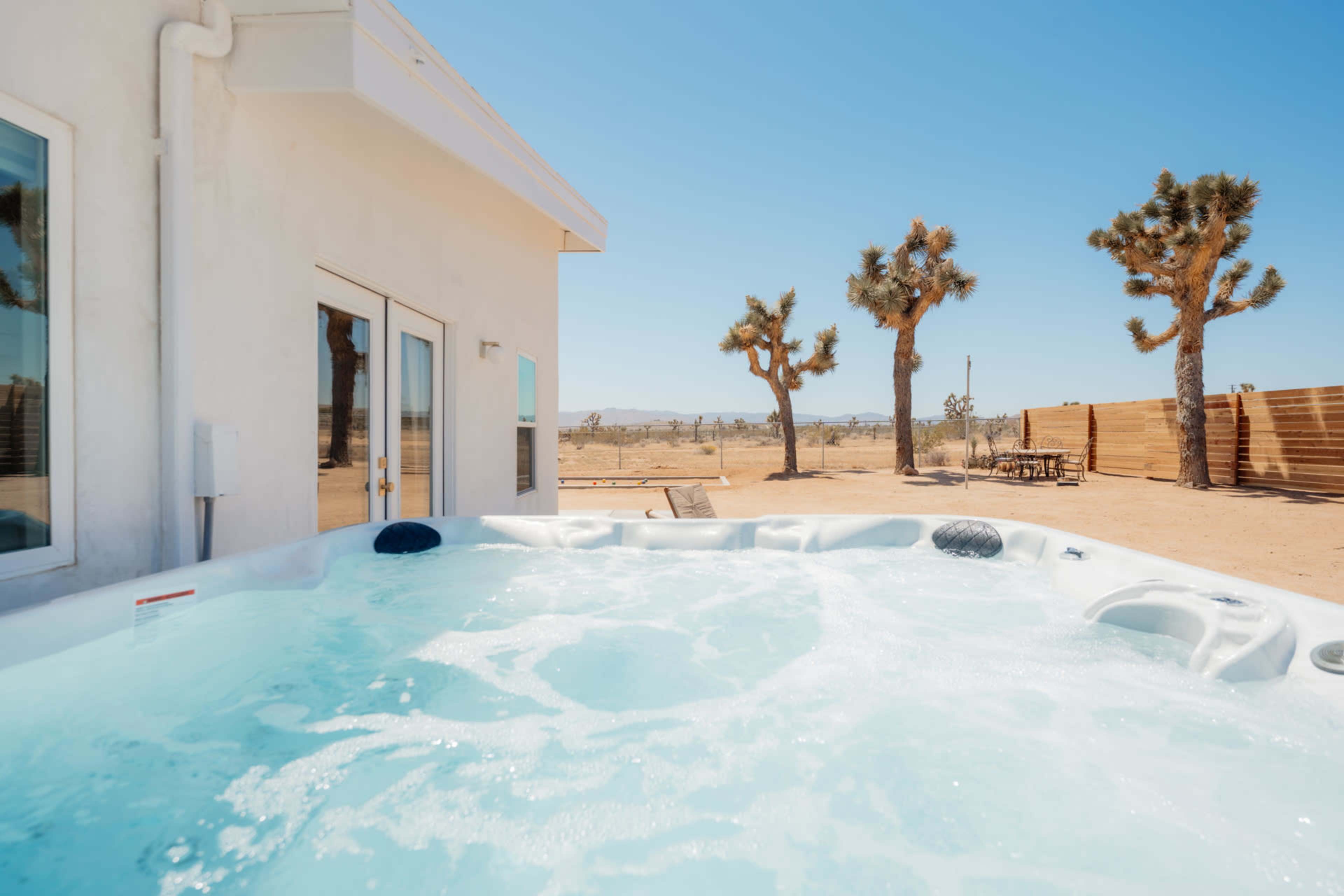 A hot tub is positioned next to a white building with Joshua trees and a picnic area visible in the background.