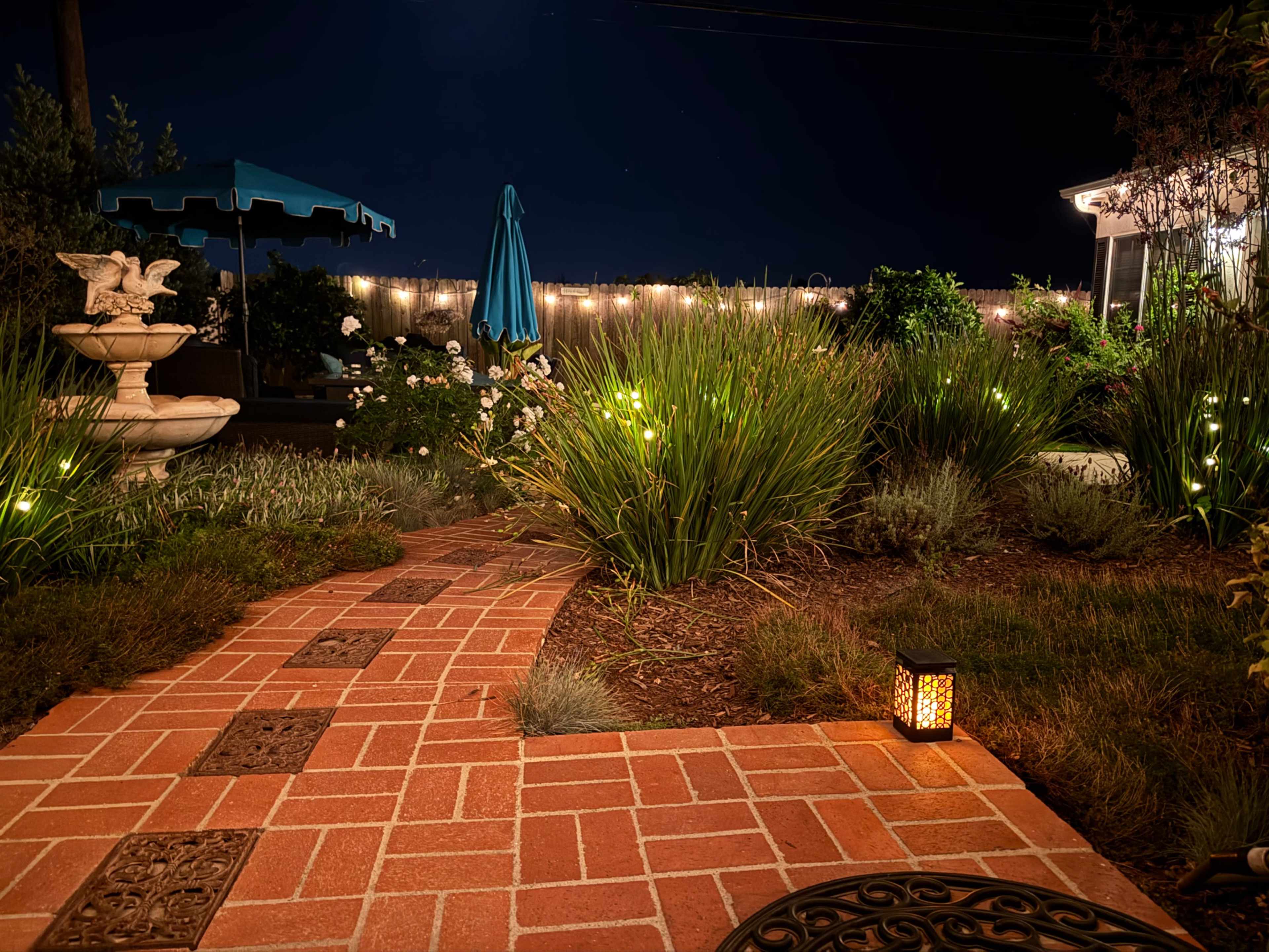 A brick pathway leads through a garden illuminated by small lights, with a fountain and a blue umbrella visible in the background.