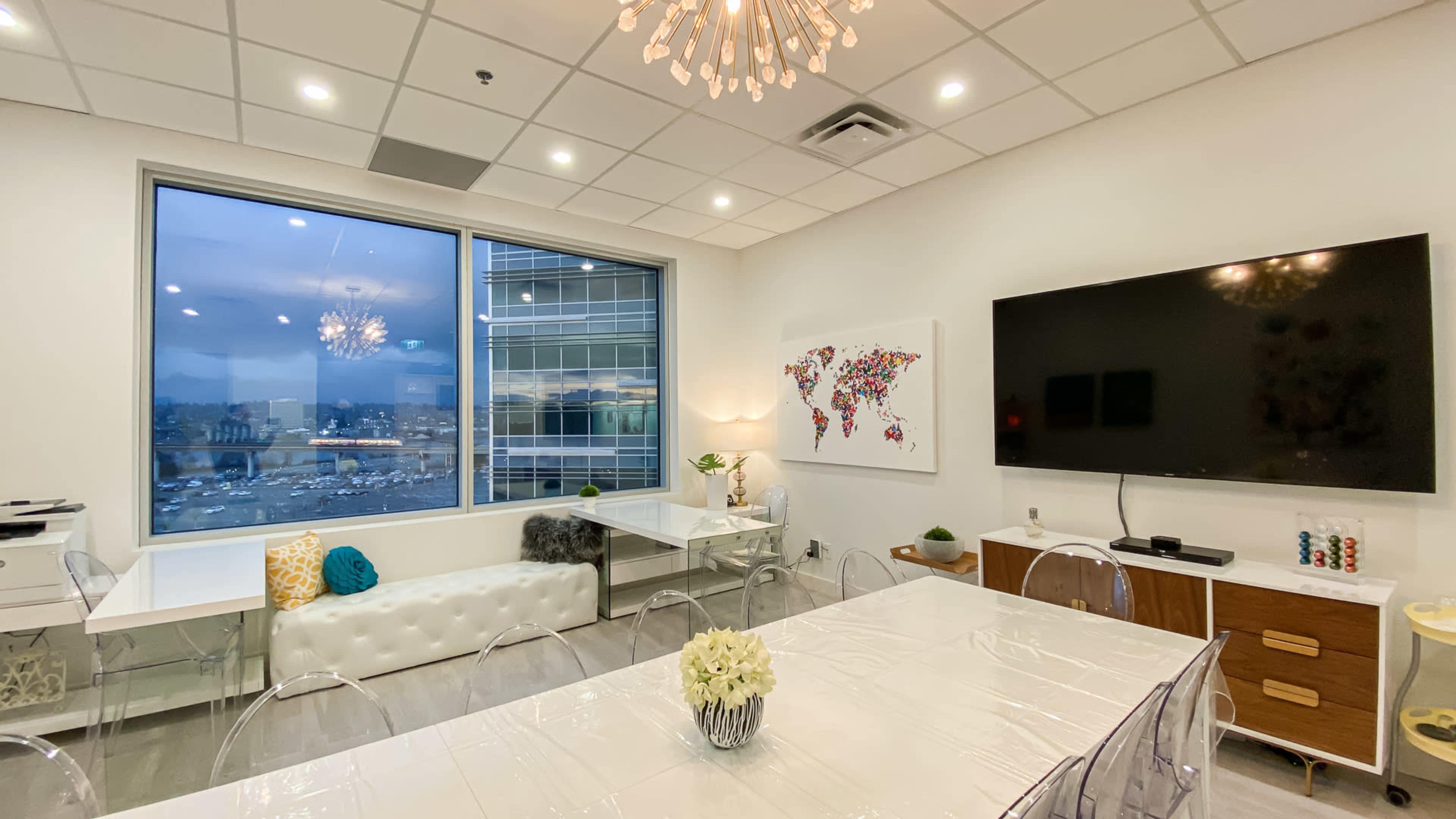 A modern conference room features a large window with a city view, a white table, transparent chairs, and a wall-mounted TV.