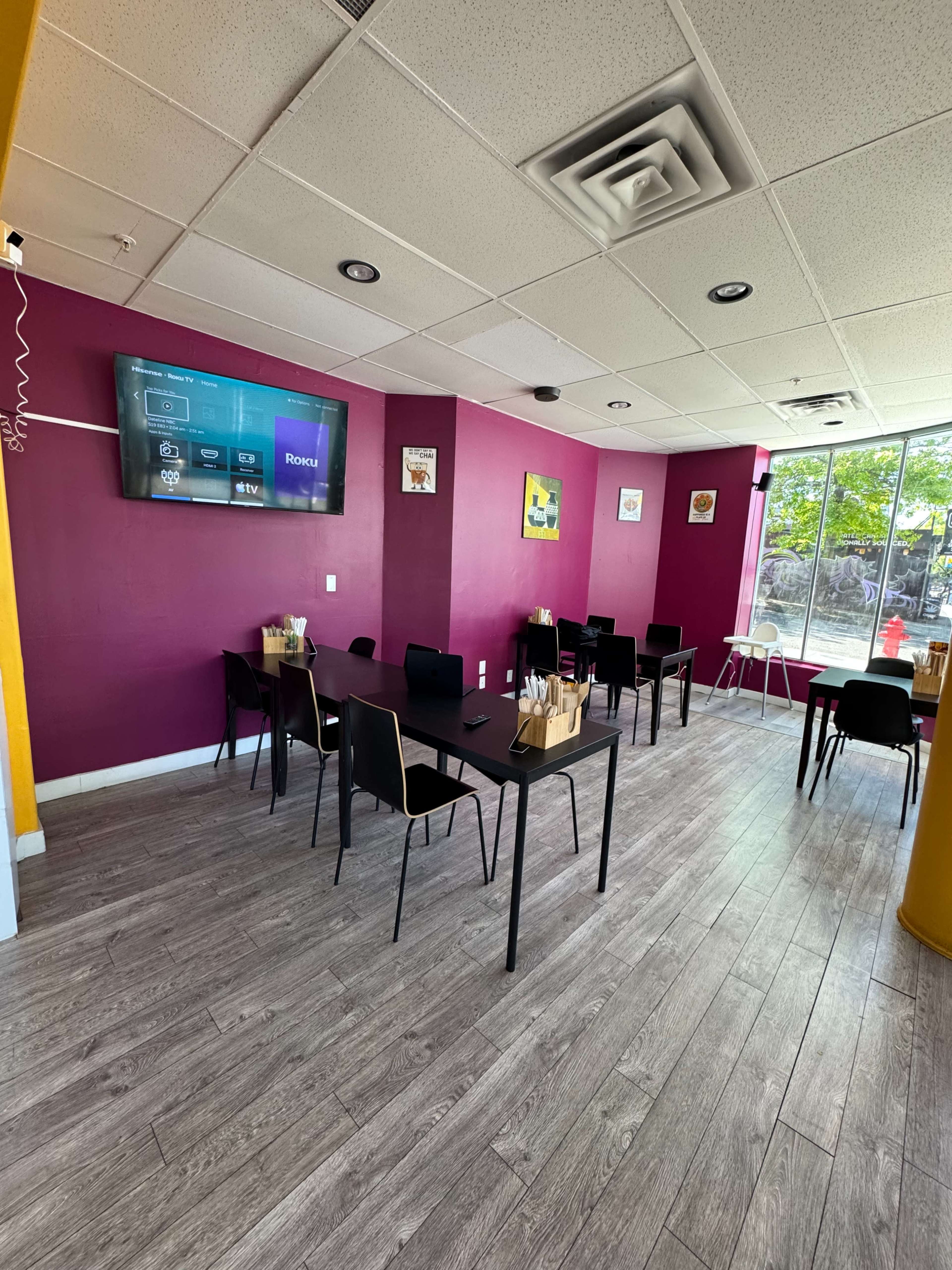 The image shows a small dining area with several tables and chairs, a wall-mounted television, and purple walls.