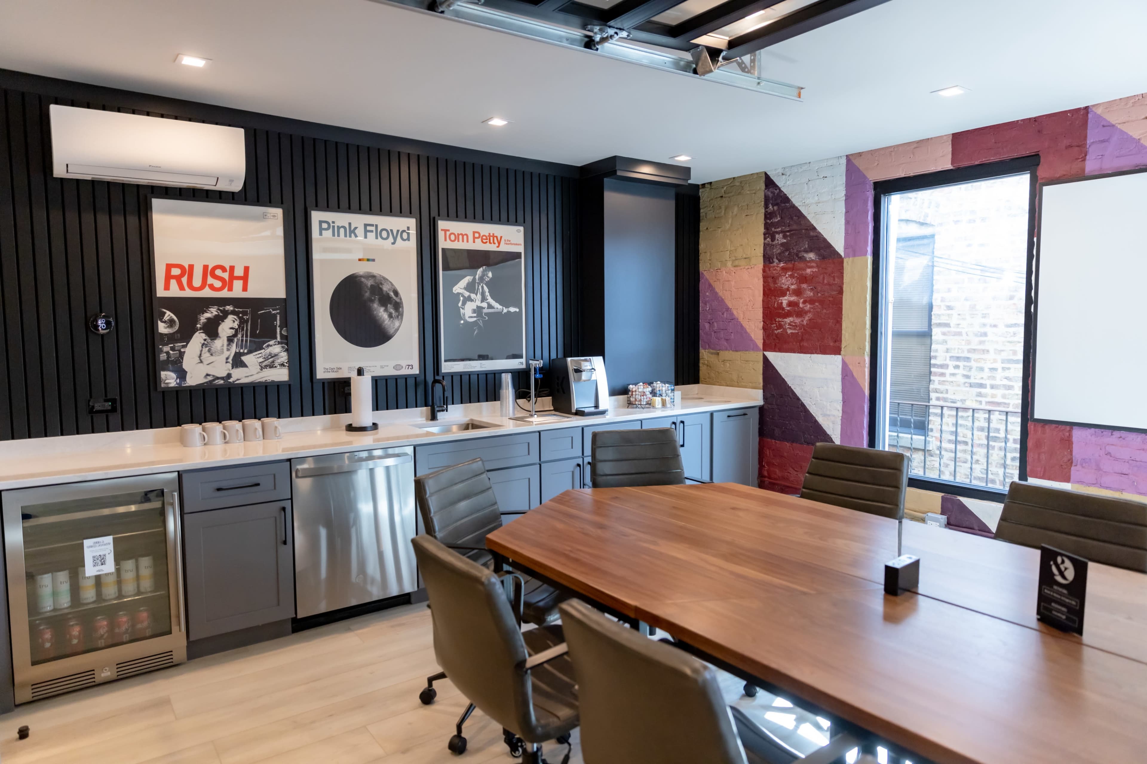 The image shows a modern conference room featuring a large wooden table, comfortable chairs, and framed music posters on the wall, with a small kitchenette area and a colorful accent wall.